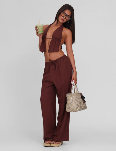 A woman wears the TIGERMIST LAINEY TOP in chocolate, a linen halter crop with adjustable ties, paired with matching wide-leg pants, sunglasses, platform sandals, a woven handbag, and holding a green drink against a light gray background.