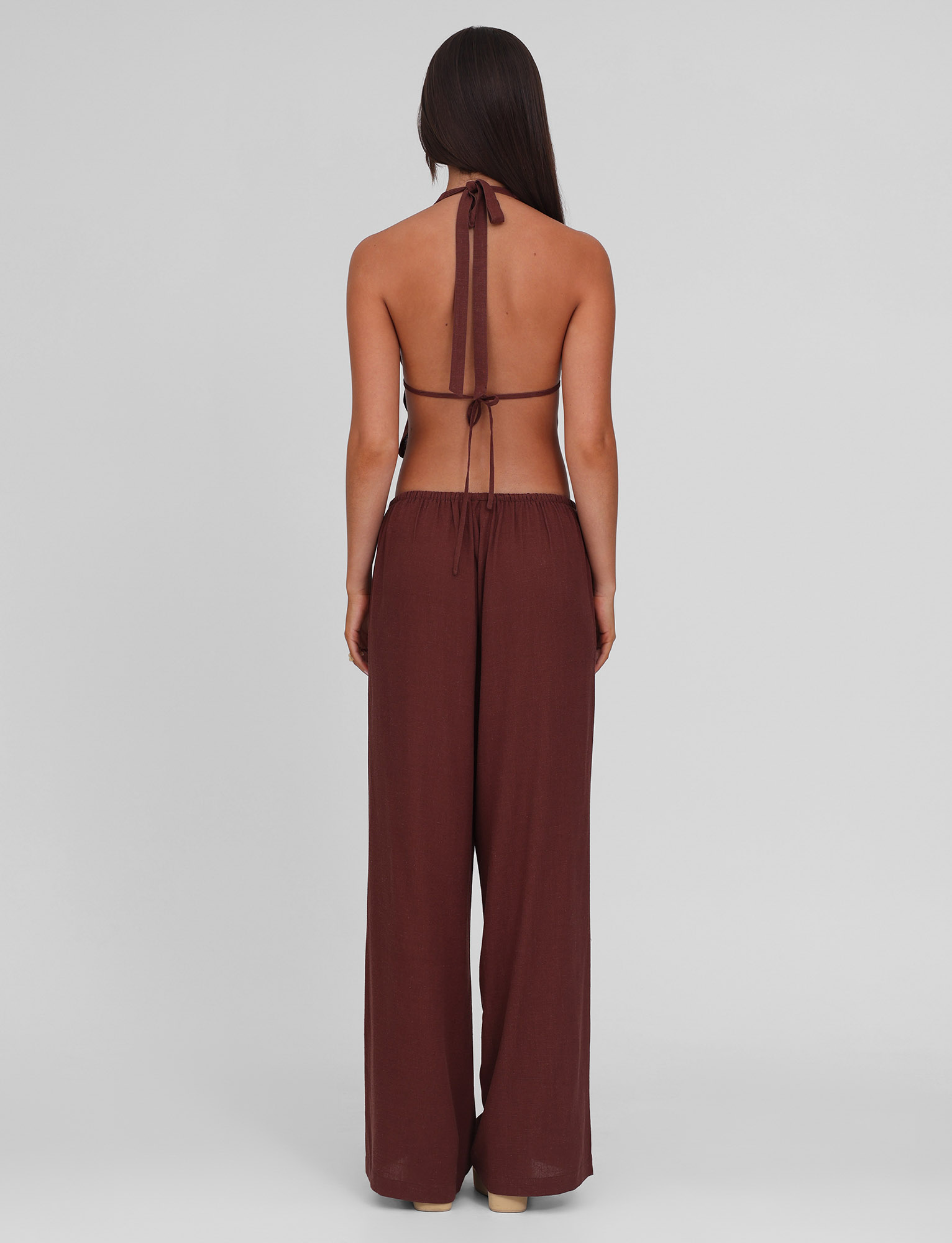 A woman with long dark hair stands facing away, wearing the TIGERMIST LAINEY PANT - CHOCOLATE, a loose, wide-leg linen style with an elasticated waist, paired with a brown halter neck backless top against a plain light gray background.