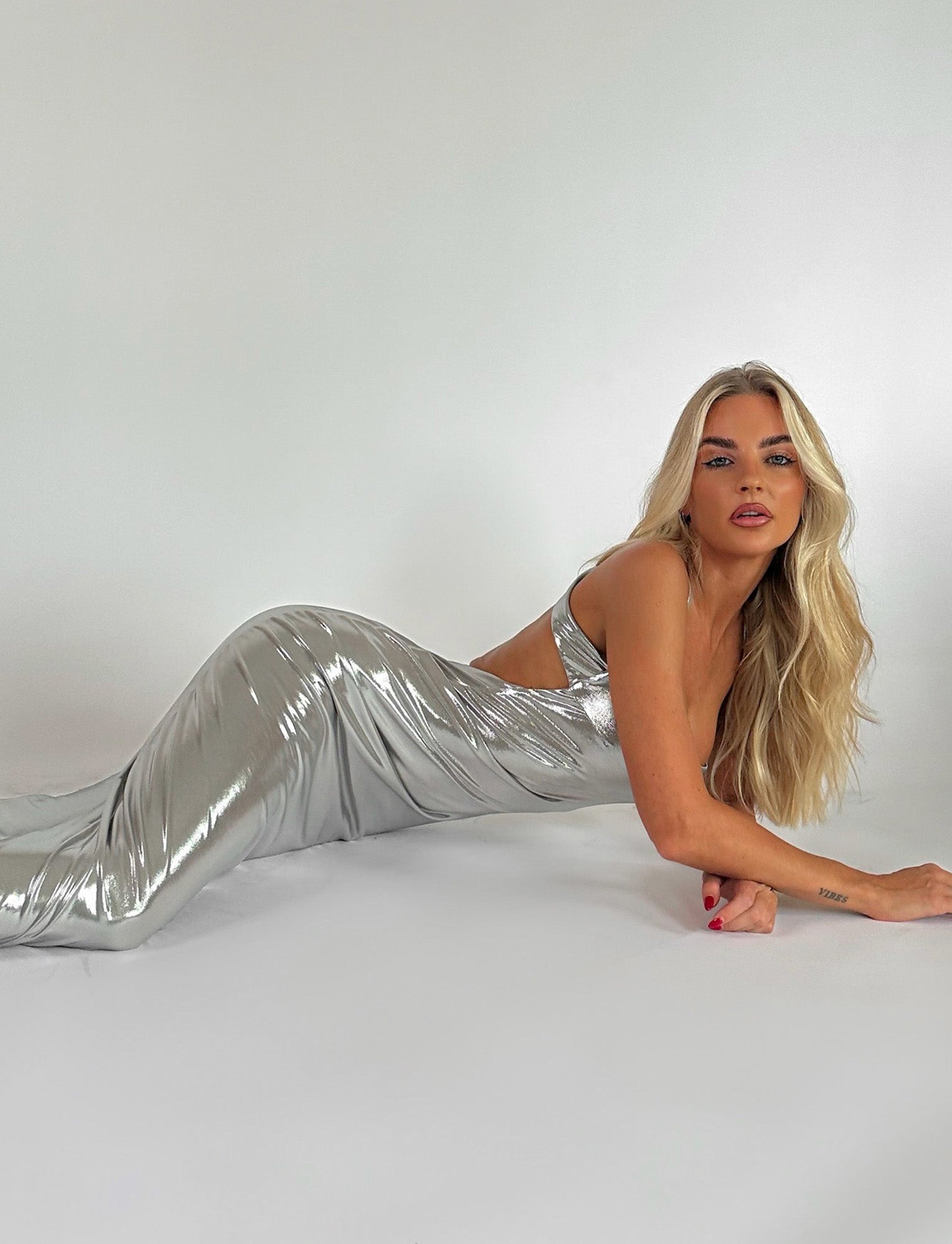 A woman with long, blonde hair is lying on her side against a plain white background. She is wearing the ELVIA MAXI DRESS - GREY : SILVER : SILVER METALLIC from TIGERMIST and looking directly at the camera with a neutral expression. She has red nail polish and a tattoo on her forearm.