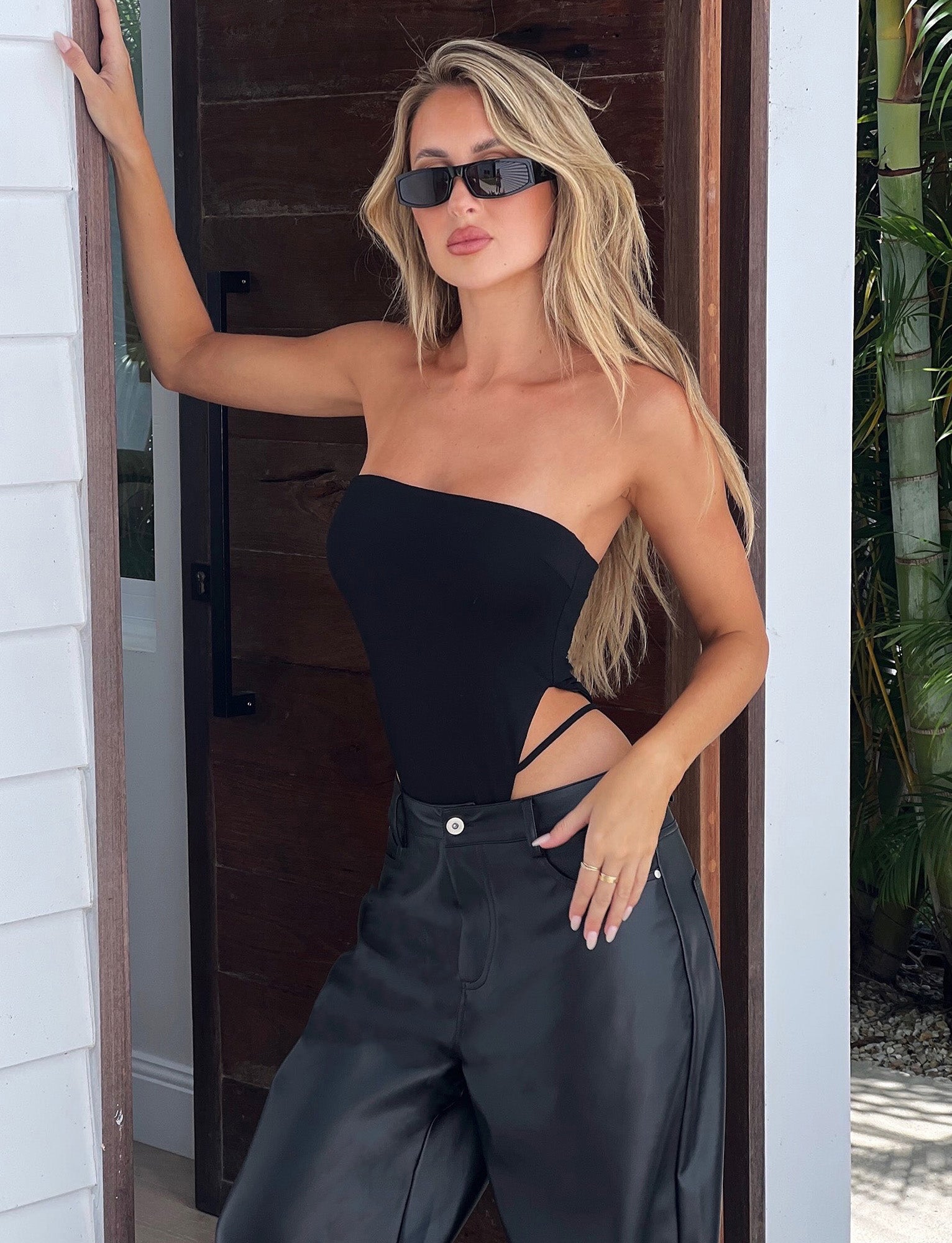 A woman with long blonde hair, wearing black sunglasses and a Tiger Mist strapless black bodysuit with side cutouts, pairs it stylishly with the TIGERMIST LELA PANT in black. She leans against a wooden door, one hand on the door frame and the other resting in her pocket.