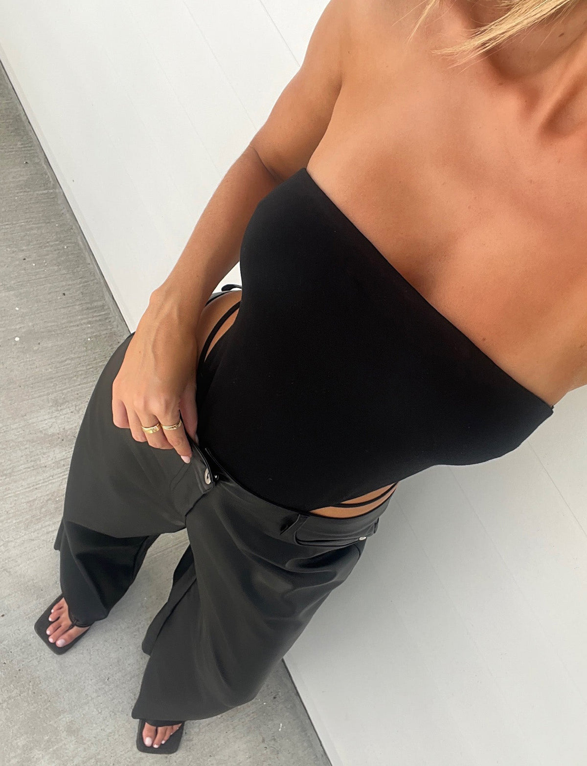 A person wearing a TIGERMIST FINIAN BODYSUIT - BLACK and dark wide-leg pants with black sandals. The background features a light-colored wall and floor. The person's face is not visible in the image.