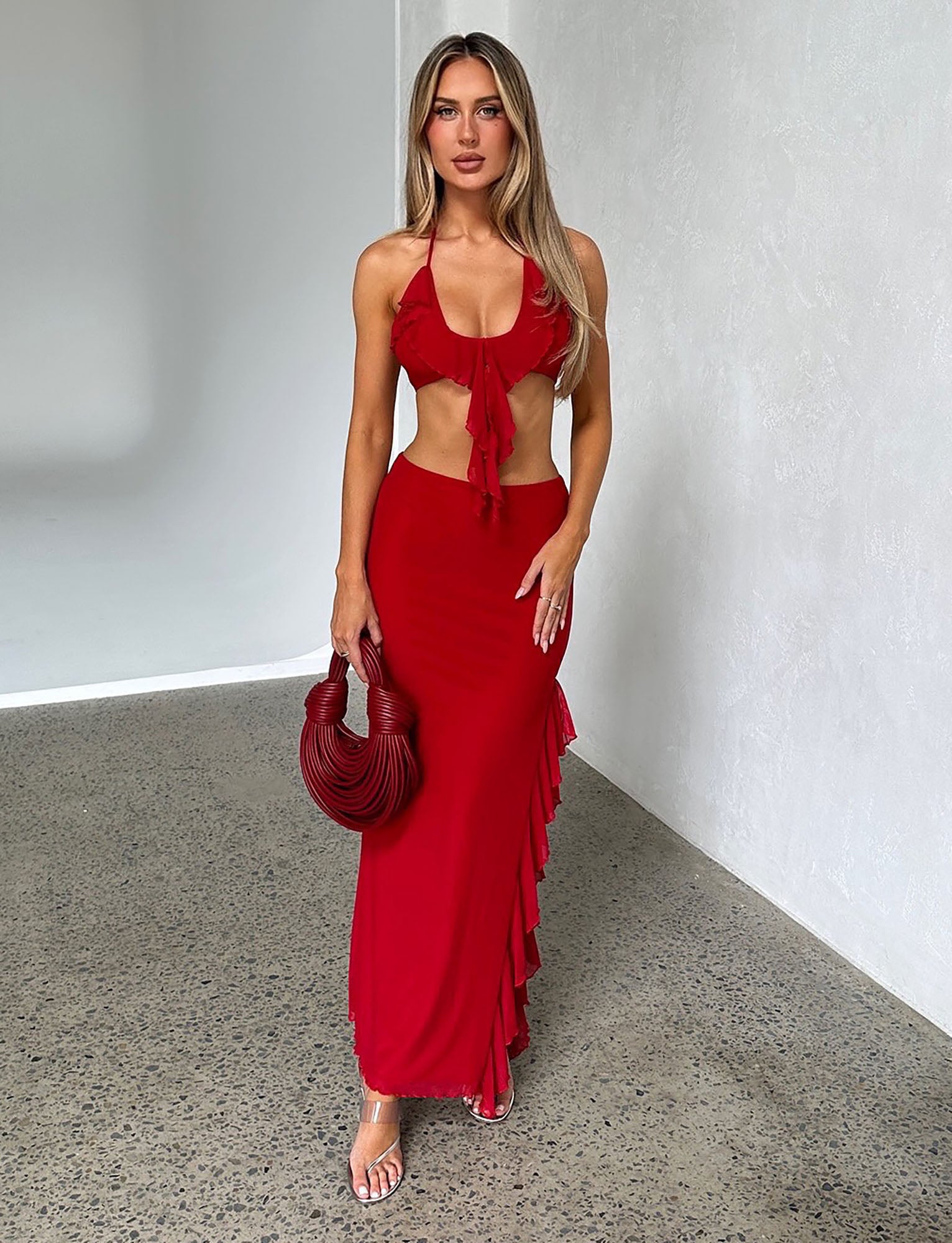 A woman stands against a light-colored wall wearing the LINETTE TOP in red and a matching long skirt with ruffle details from TIGERMIST. She holds a red, textured handbag and wears transparent high heels. Her long hair is styled in loose waves, and she poses with a neutral expression.