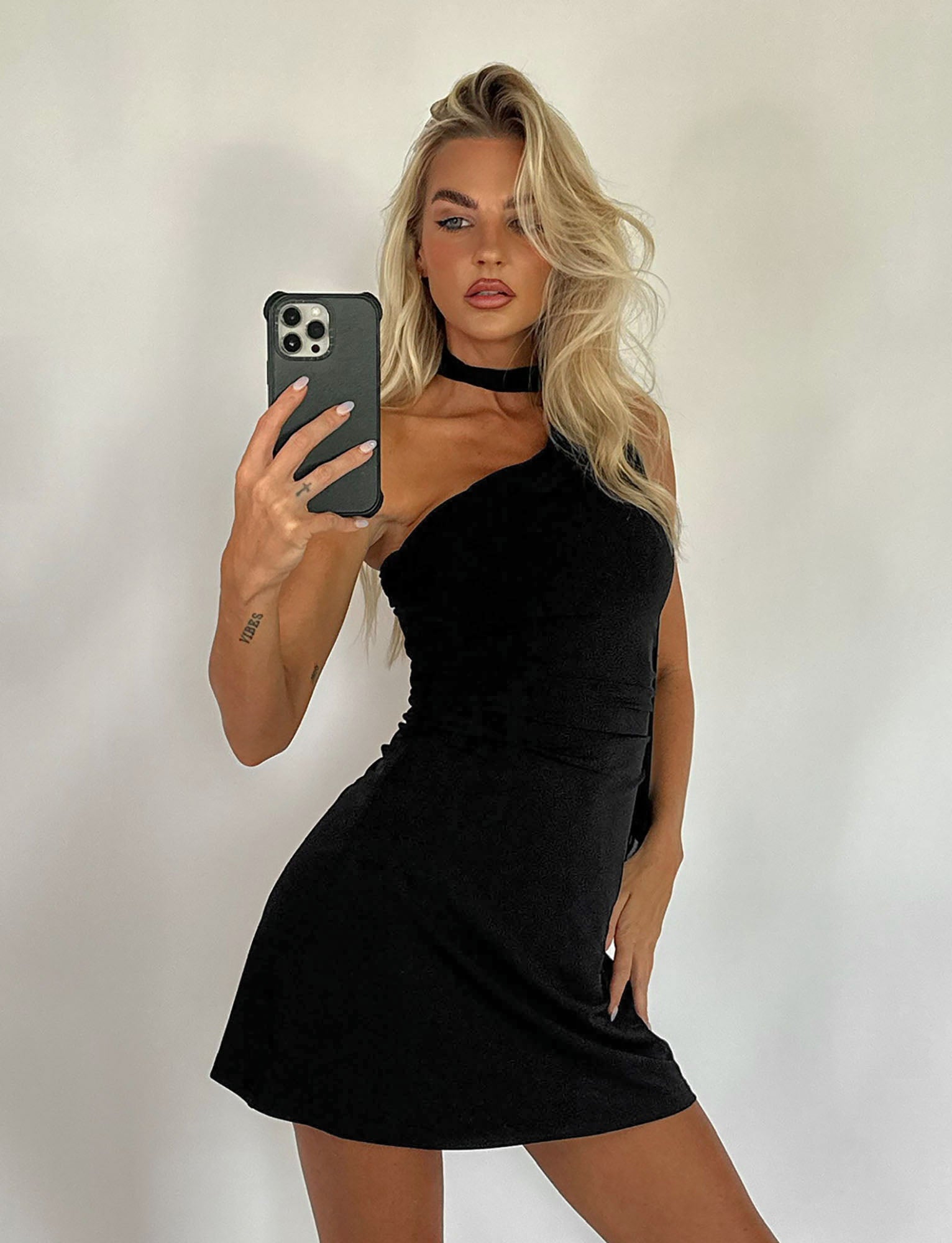 A person with long, blonde hair stands against a plain background, holding a smartphone and taking a mirror selfie. They are wearing the ALLIRA DRESS - BLACK from TIGERMIST, which features a one-shoulder design and a choker neckline. A tattoo is visible on their right arm.