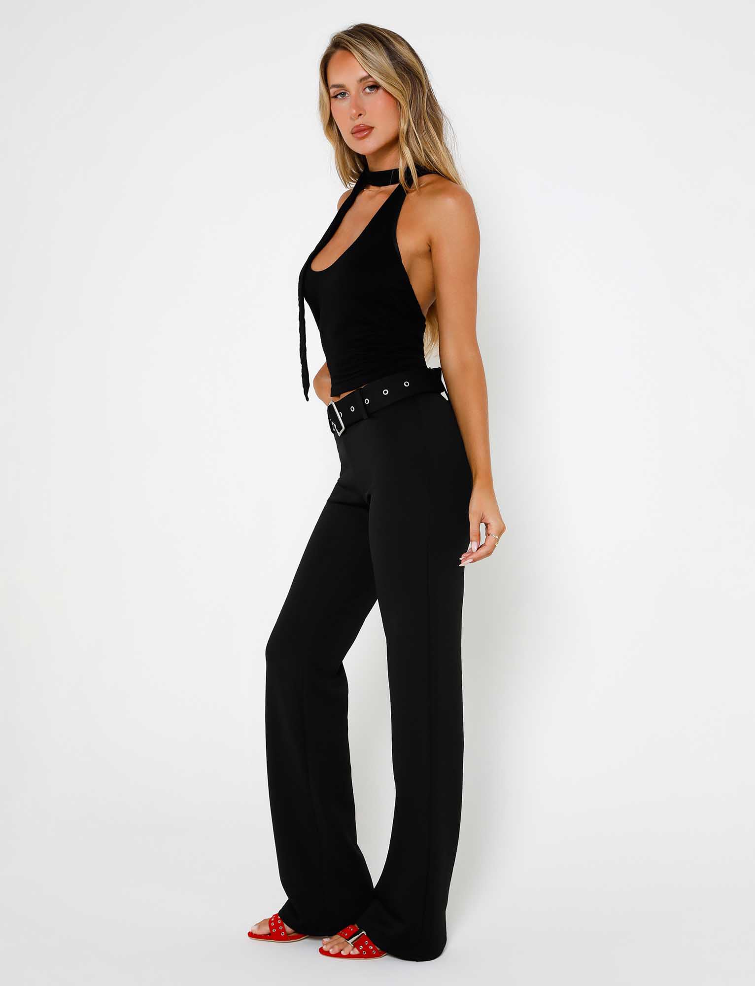 A woman with long blonde hair stands against a white background, dressed in the HADLEY PANT - BLACK from TIGERMIST. She pairs it with a black halter top and a matching belt, completing her look with red shoes and striking a confident pose, facing the camera with her body slightly angled to the side.