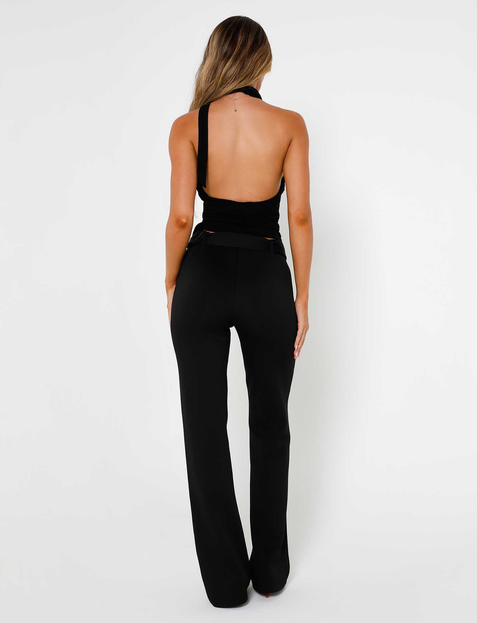 A woman is standing with her back to the camera, wearing a black, one-shoulder, backless top paired with HADLEY PANT - BLACK from TIGERMIST. Her long hair cascades down her back against a simple white backdrop.