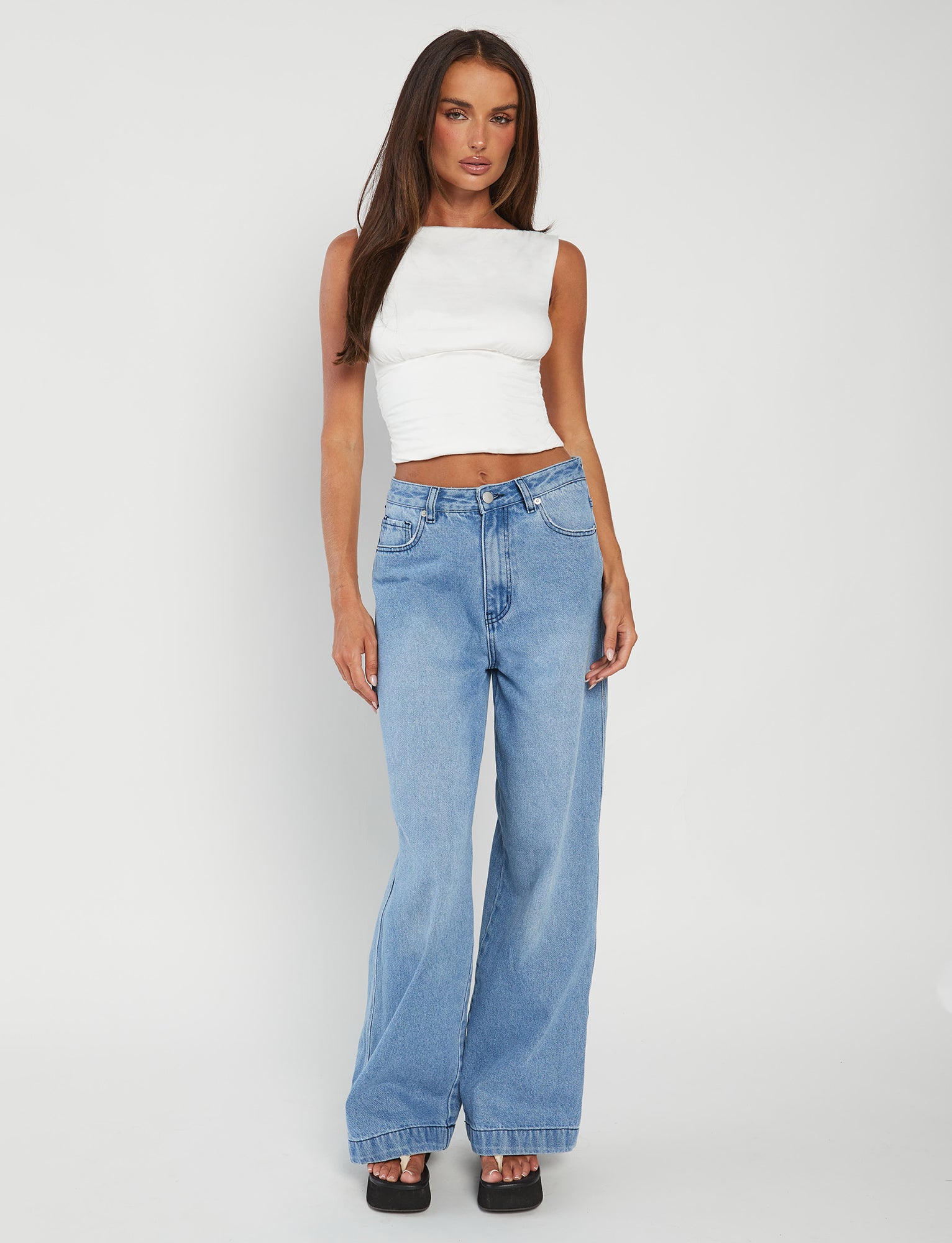 A woman stands against a plain white background, modeling the GWYN TOP - WHITE from TIGERMIST and high-waisted, loose-fitting blue jeans. She has long, straight hair and is wearing black platform shoes. With her arms relaxed by her sides, she looks directly at the camera.