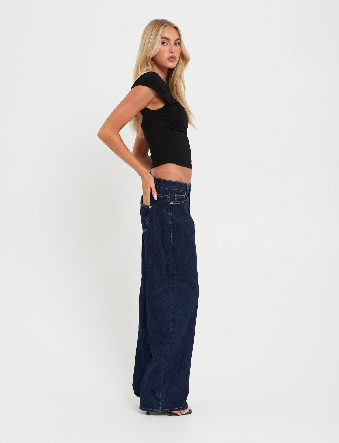 A woman with long blonde hair wearing the RYLAN TOP - BLACK from TIGERMIST and loose dark blue jeans stands sideways against a plain white background. She has one hand in her pocket and is looking towards the camera.