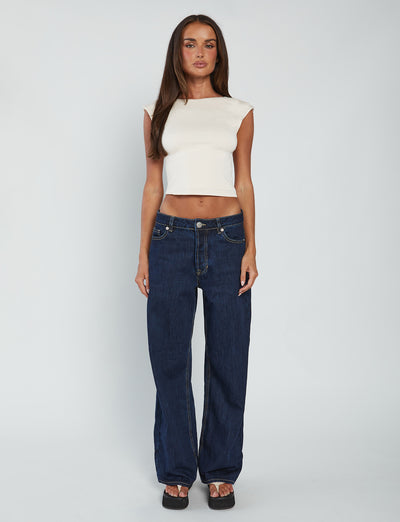 A woman stands against a plain white background. She has long, straight brown hair and is wearing a sleeveless, form-fitting Tigermist Rylan Top in Beige : Sand, dark blue jeans that are slightly loose with visible stitching, and black platform sandals. Her arms are relaxed by her sides.