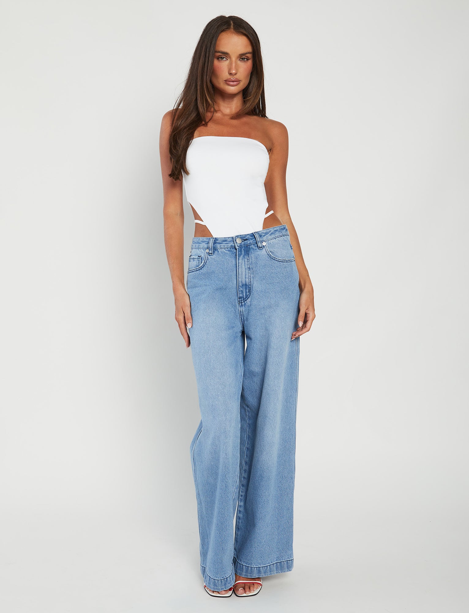 A woman stands against a plain white background, wearing the FINIAN BODYSUIT in white from TIGERMIST and high-waisted, light blue wide-leg jeans. She has long, straight hair and is posing with one hand on her side. She is wearing white high-heeled shoes.