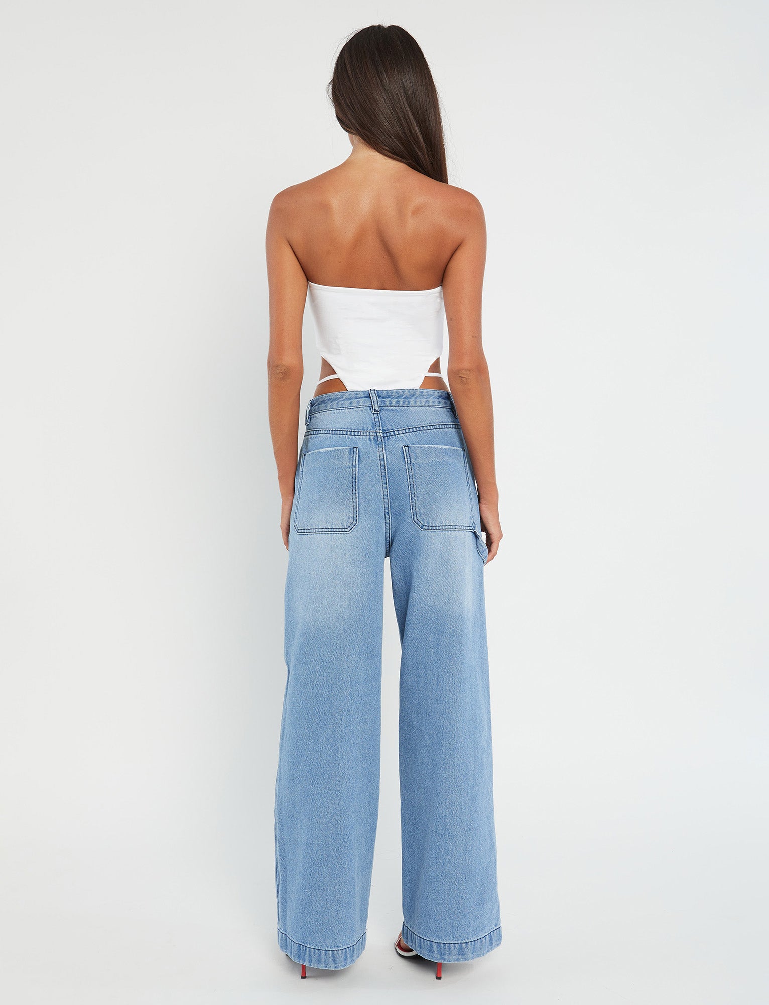 A person with long dark hair stands with their back to the camera against a plain white background. They are wearing a chic Tiger Mist FINIAN BODYSUIT in white paired with high-waisted, light blue wide-leg jeans.