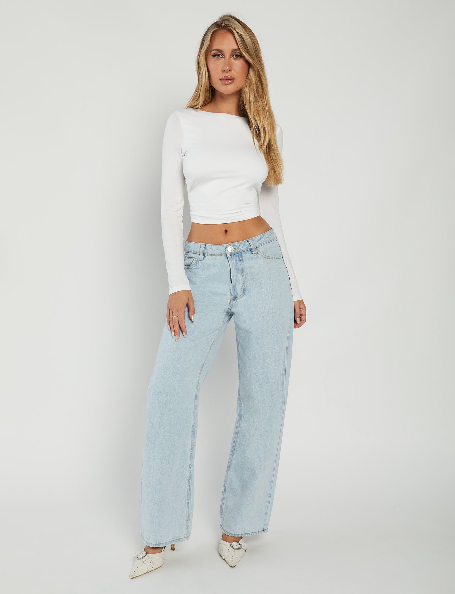 A woman stands against a plain white background, wearing the QUINNELL TOP - WHITE from TIGERMIST, a long-sleeved, fitted white crop top. She pairs it with light blue high-waisted wide-leg jeans and is styled with long, wavy blonde hair. She completes her look with white pointed-toe shoes and poses gracefully with her left hand resting on her hip.