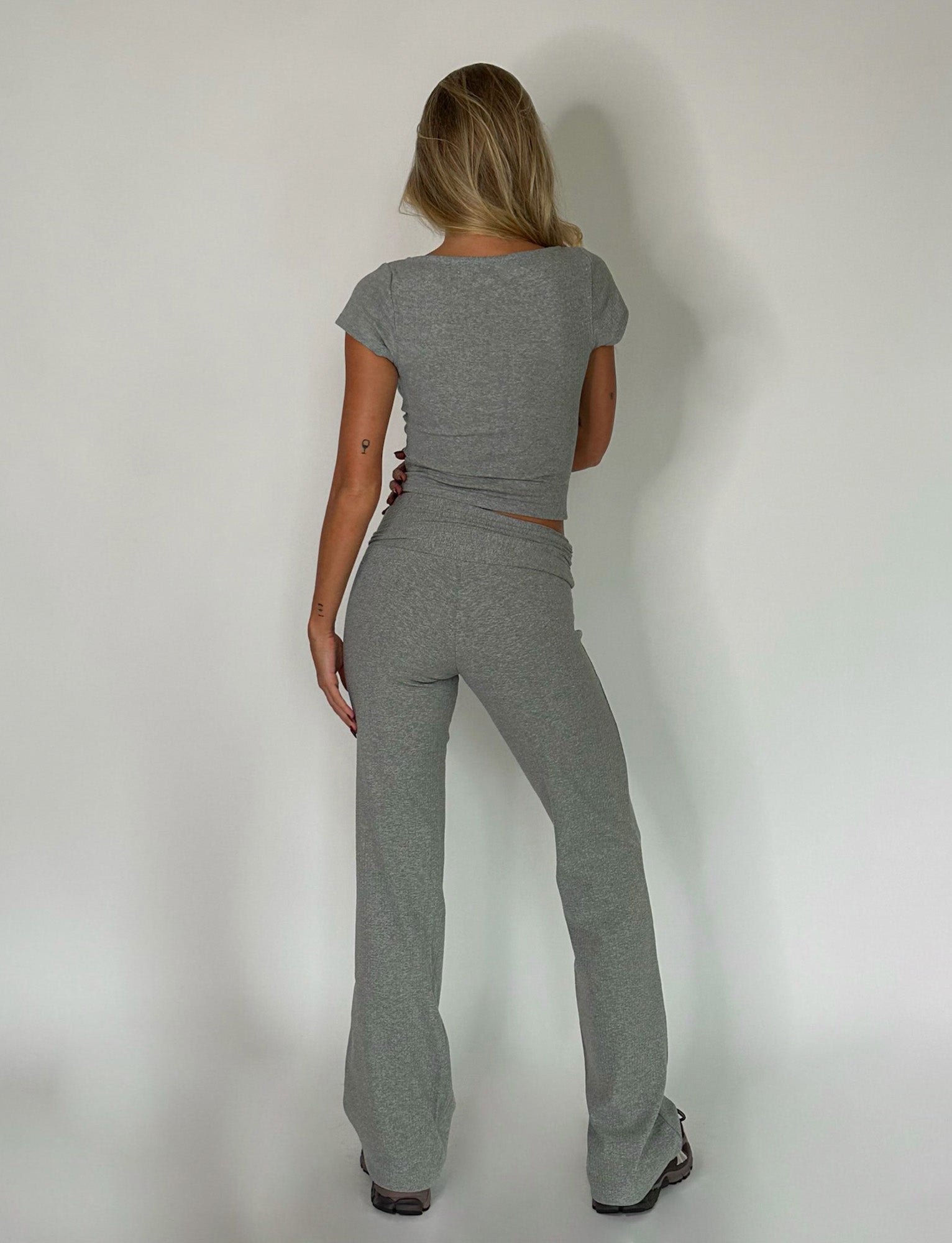 A person with long blonde hair stands facing a white wall, their back to the camera. They are wearing a fitted Dove Grey short-sleeve Kavat Top and matching gray pants from Tiger Mist. The lighting is soft, creating subtle shadows on the wall.