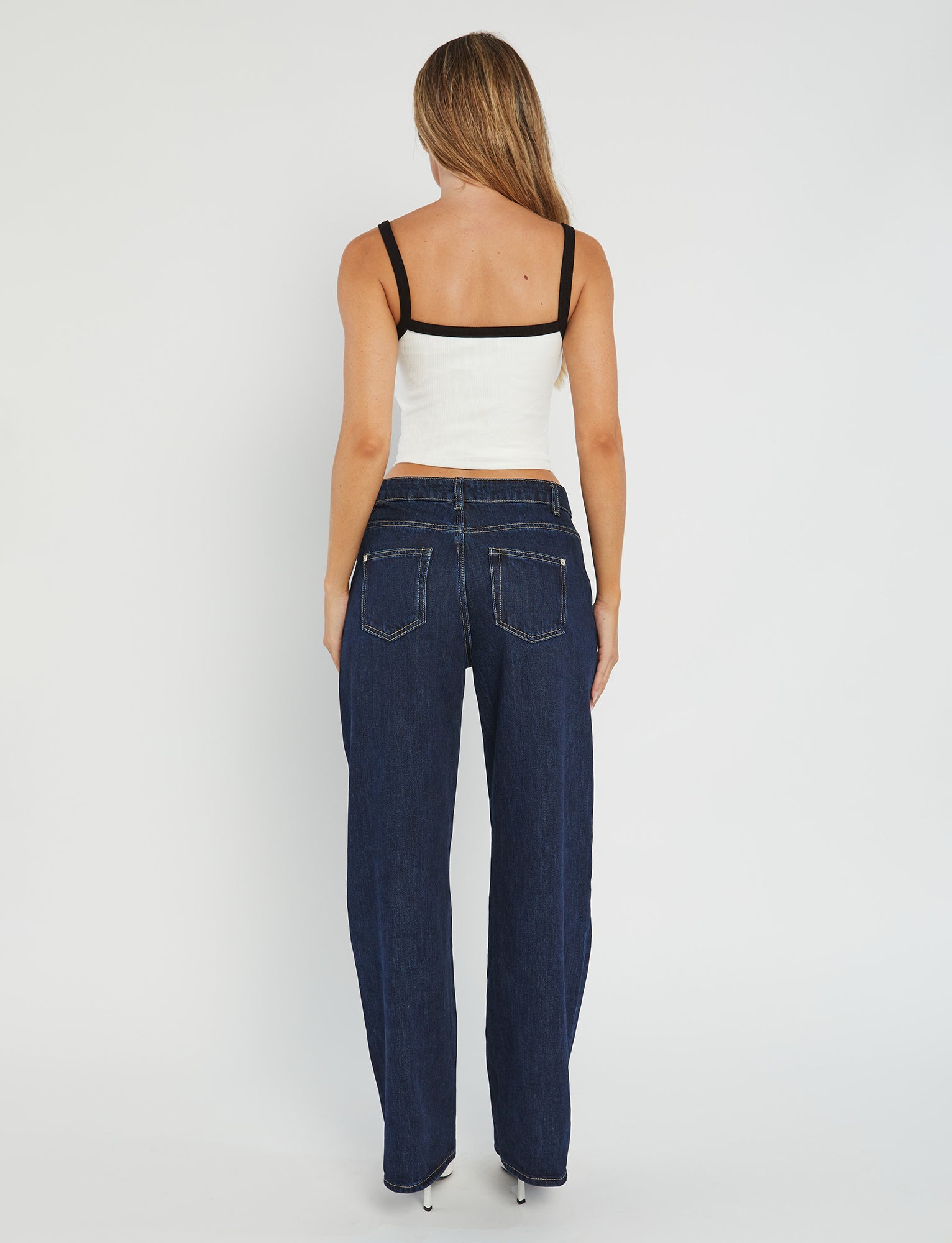 A woman with long hair stands facing away, modeling the NIXIE TOP - WHITE from TIGERMIST, paired with dark blue jeans. The scene is set against a plain white background.
