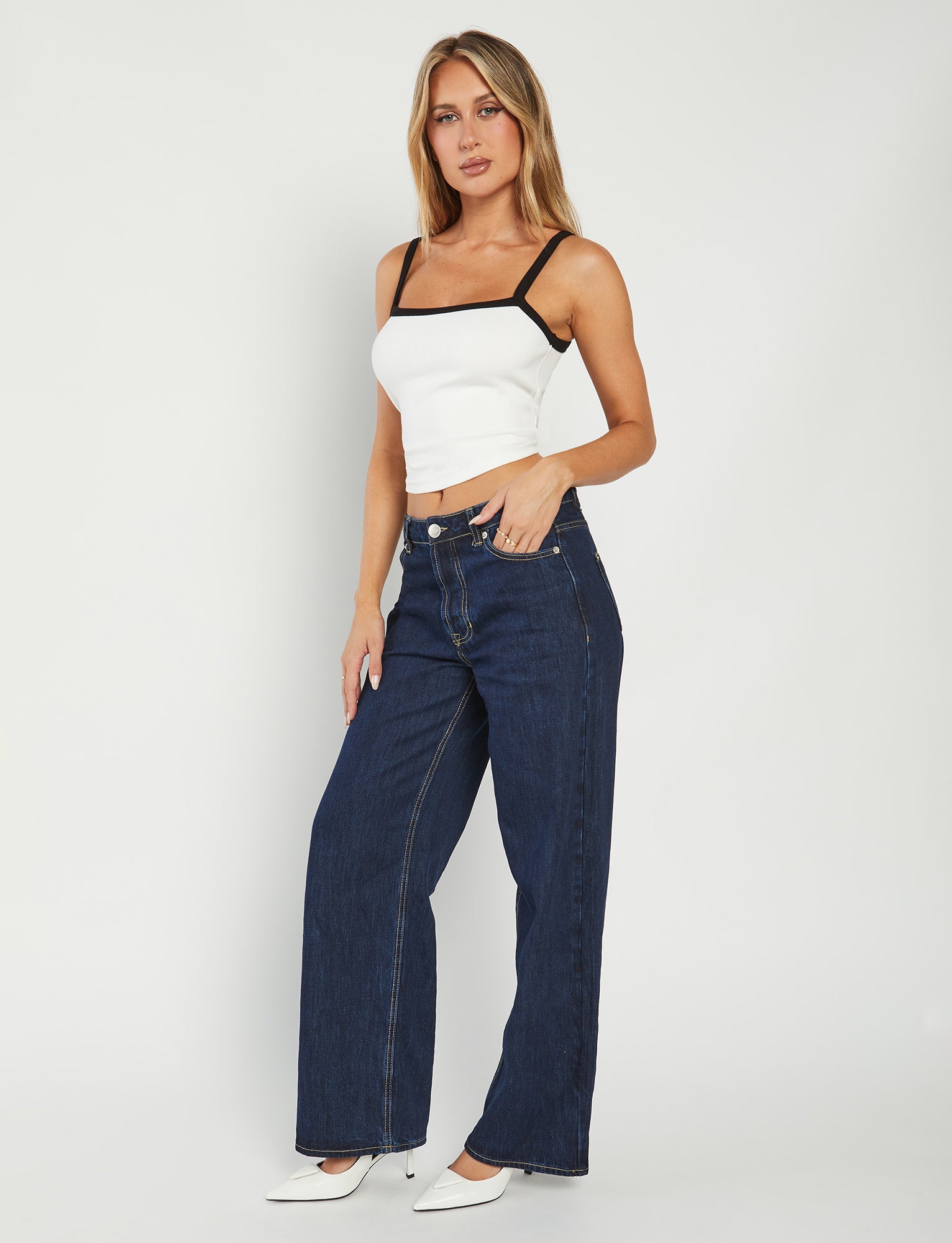 A woman stands against a plain white background wearing a chic TIGERMIST NIXIE TOP in off white with black straps, high-waisted dark blue jeans, and matching white heels. Her right hand rests on her hip while her left arm hangs down by her side. Her hair is styled in loose waves.