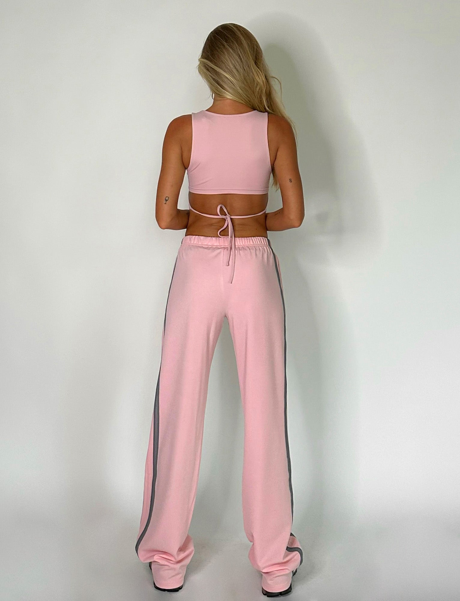 A person with long blonde hair is facing away from the camera, wearing the TIGERMIST LERO TOP in baby pink and matching Tiger Mist pink sweatpants with white stripes down the sides. They are standing against a plain white background.