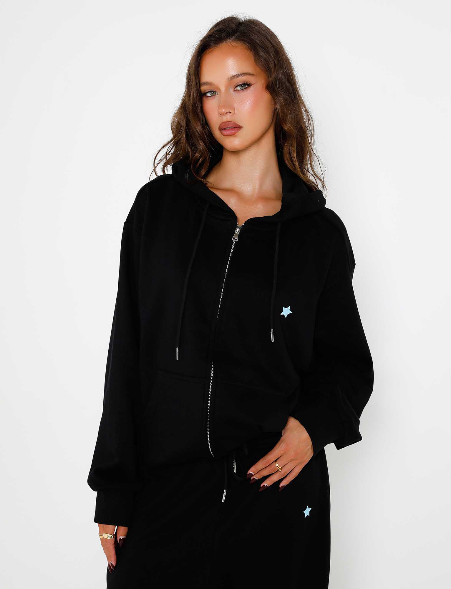 A woman with long, wavy hair is wearing the ALISH HOODIE - BLACK by TIGERMIST, which features a small blue star on one side. She stands against a plain white background, looking directly at the camera with a neutral expression while her left hand holds the edge of the hoodie.