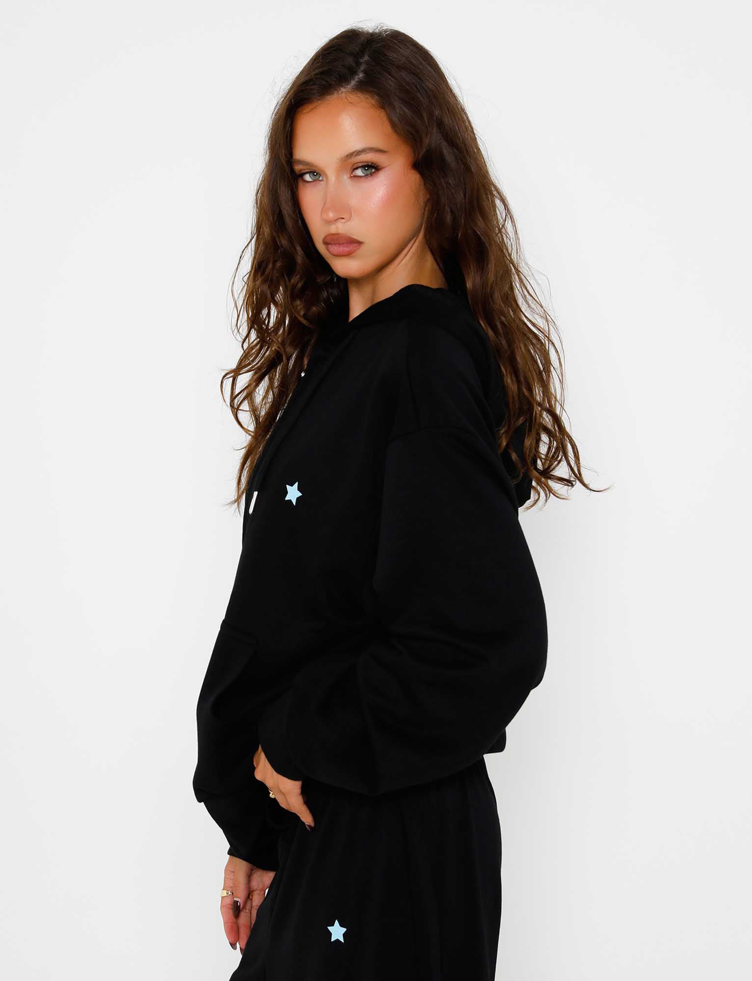 A woman with long brown hair is posing against a plain white background. She is wearing the ALISH HOODIE - BLACK by TIGERMIST and matching pants, both featuring small blue star details. She gazes into the camera with a neutral expression, her hands resting at her sides.