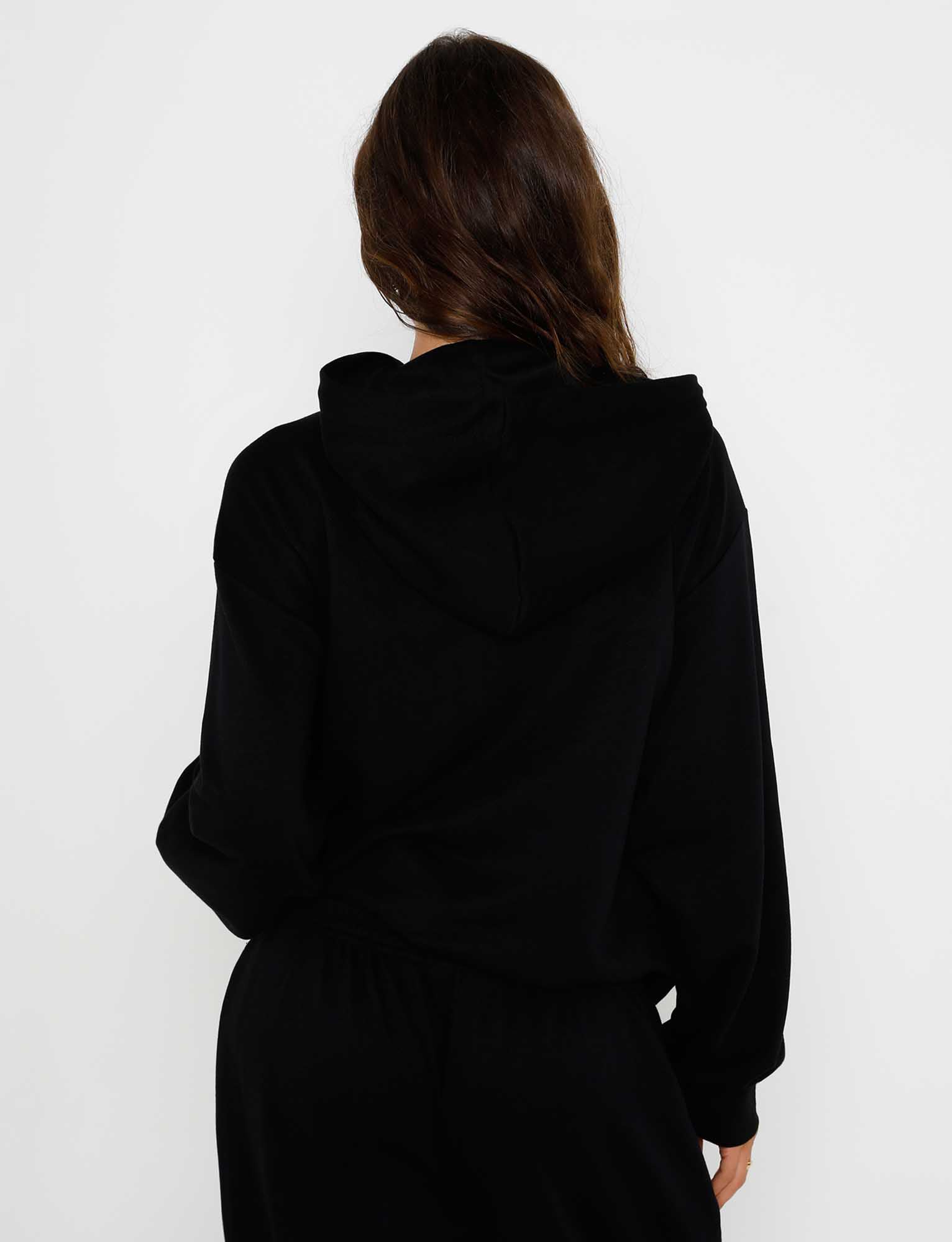 An individual with long, wavy hair is shown from the back, wearing an ALISH HOODIE in black by TIGERMIST along with black pants. The plain white background accentuates the dark clothing.