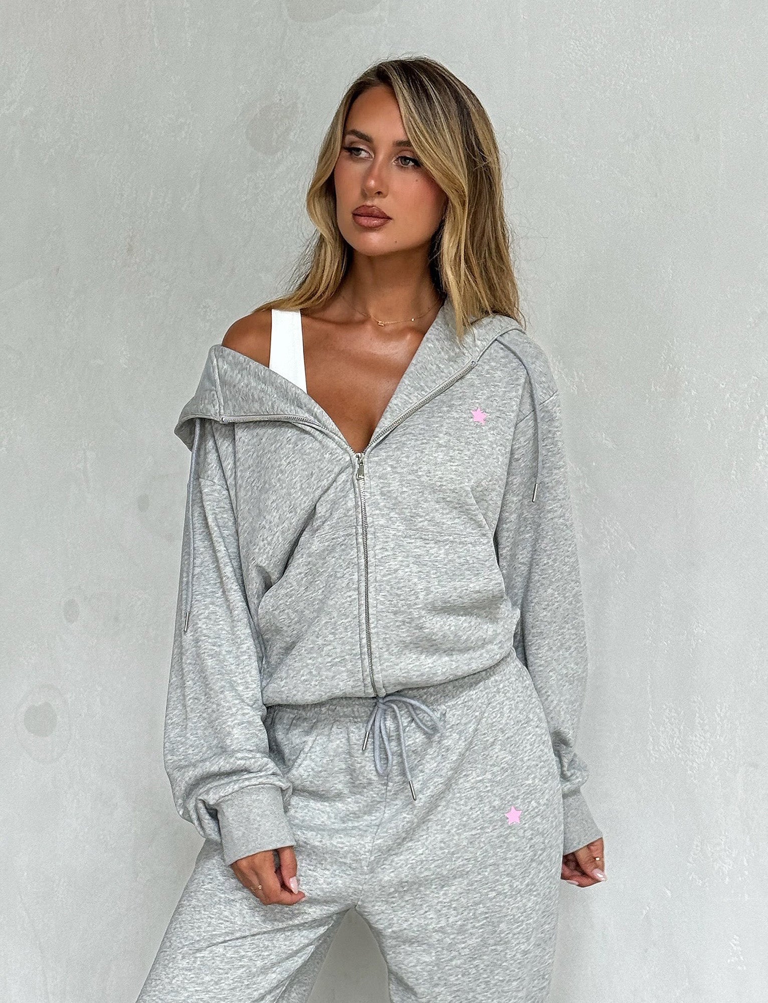 A person stands against a light grey wall, wearing a stylish TigerMist ALISH HOODIE - GREY : GREY MARLE with the zipper partially open, revealing a white top underneath. Their blonde hair is loose, and they have a relaxed expression.