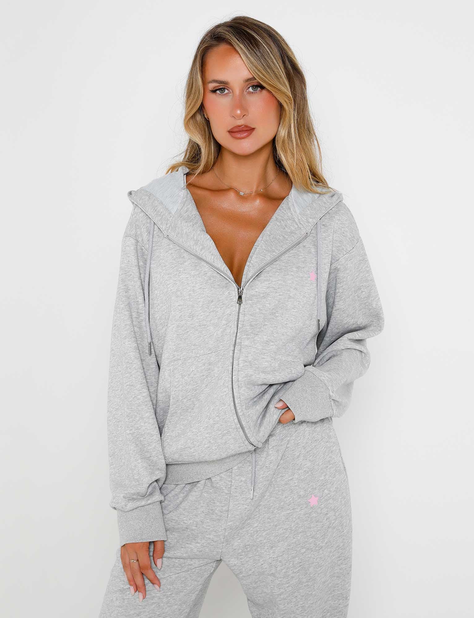 A woman with long blonde hair is wearing a light grey Alish Hoodie and matching sweatpants from TigerMist. She stands against a plain white background with her left hand in her pocket and her right arm resting casually by her side. She has a neutral expression on her face.