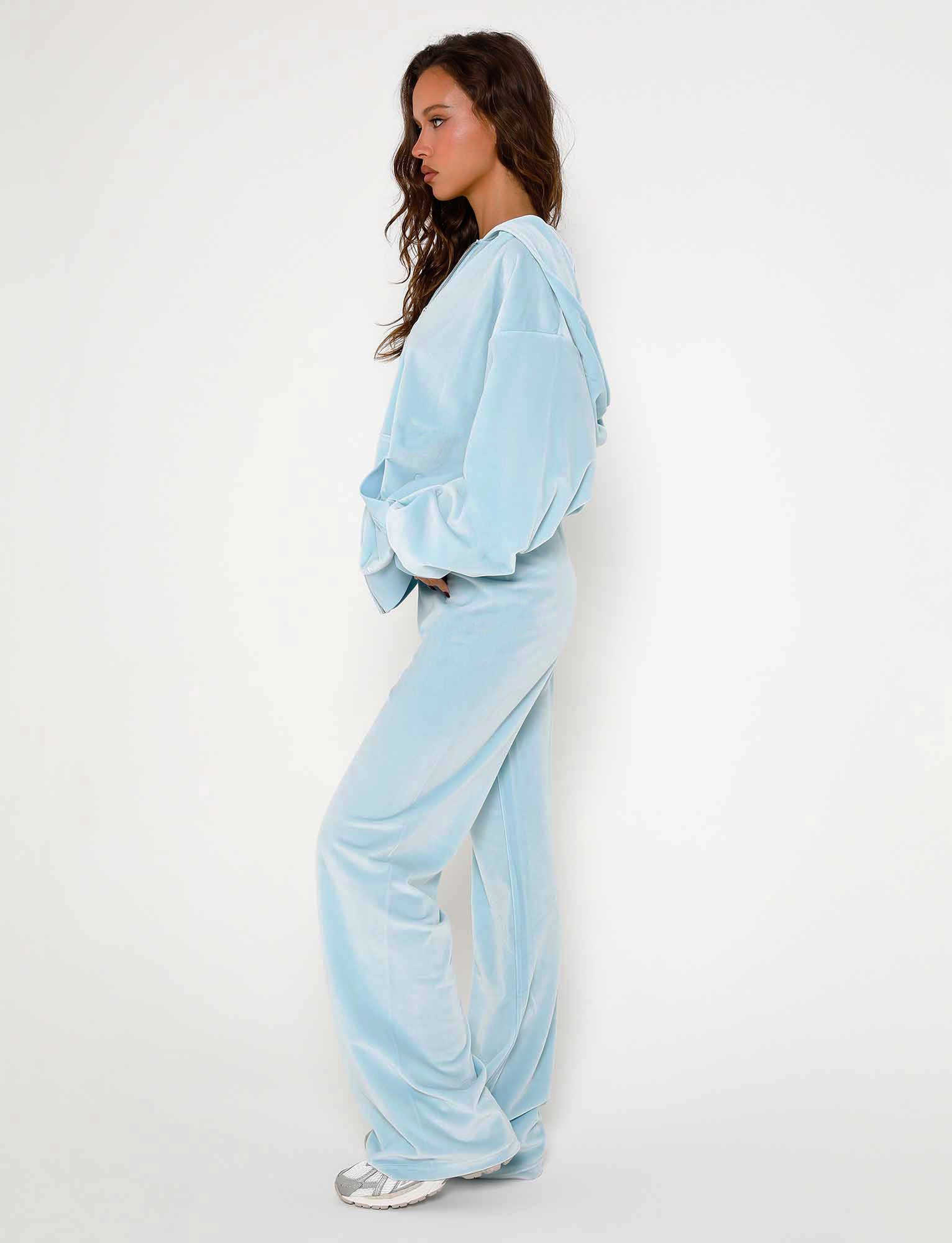 A woman with long, wavy hair stands in profile, facing left. She wears a light blue, hooded velour tracksuit from TigerMist featuring the ERICKA PANT in baby blue with wide-leg styling and holds her hands in her pockets. She completes the outfit with white sneakers. The background is a plain white wall.