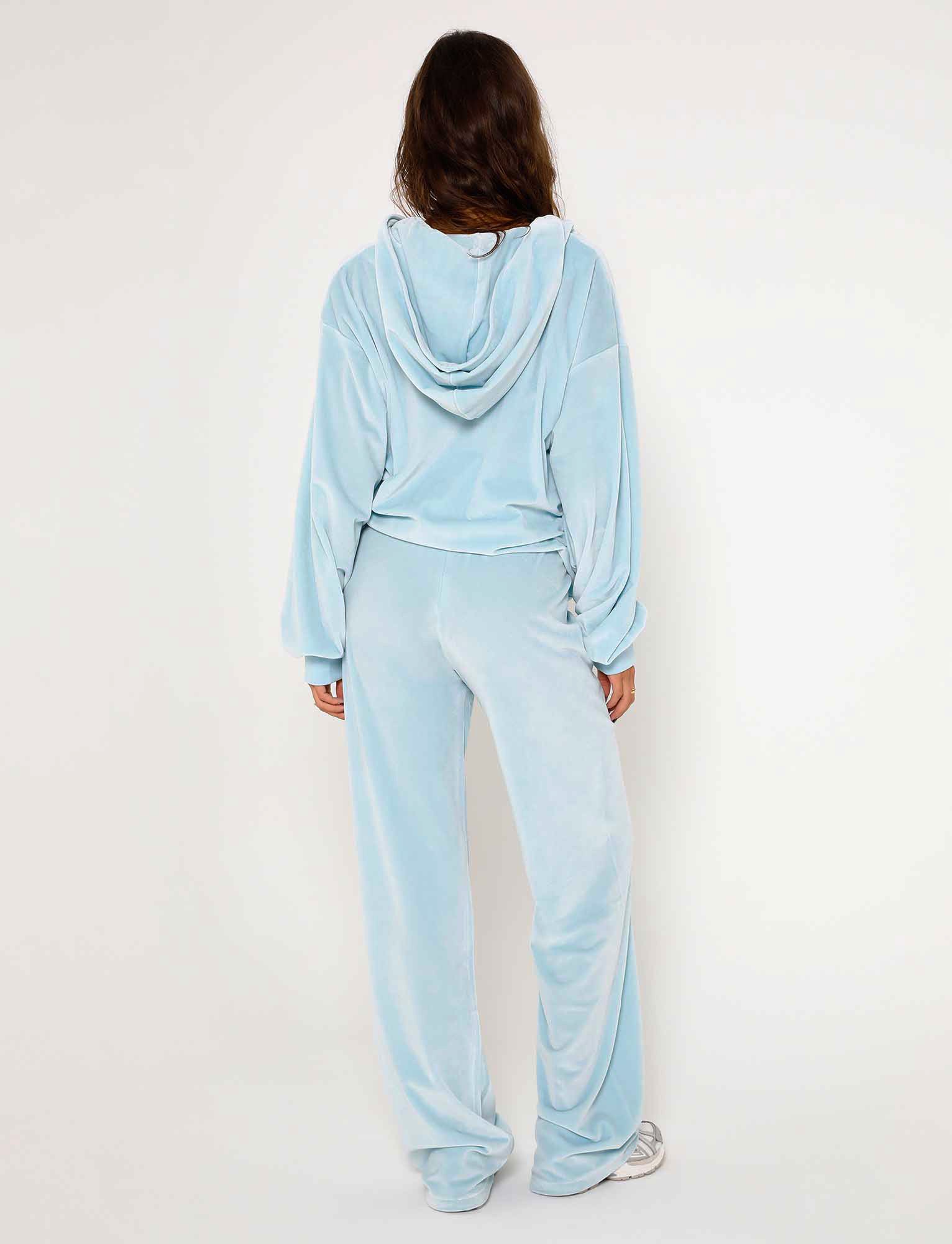 A person with long hair is standing with their back to the camera, dressed in a light blue TIGERMIST velour tracksuit featuring the ERICKA PANT - BLUE : BABY BLUE, complete with a hood and long sleeves. They are also wearing white sneakers. The background is plain and light-colored.