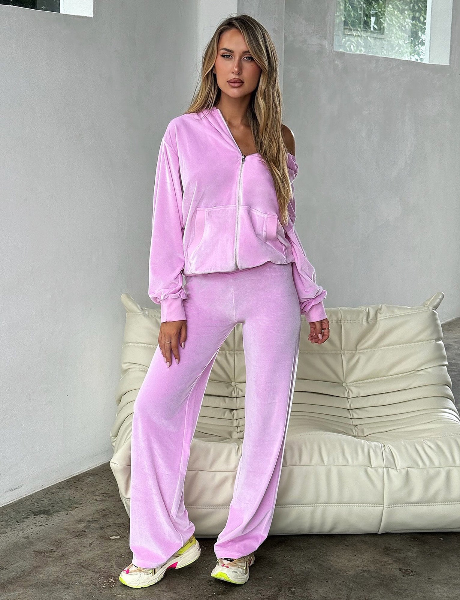 A woman with long blonde hair is standing indoors wearing the BABY PINK ERICKA PANT from TIGERMIST. She has one hand in her pocket and is leaning slightly against a cushioned beige chair, paired perfectly with white and neon yellow sneakers.