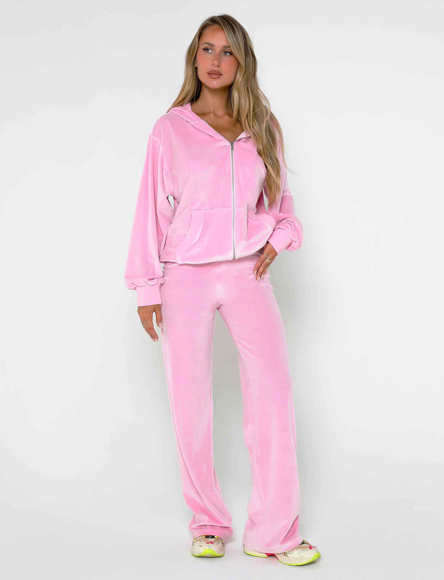 A woman stands in front of a plain white background, wearing the TIGERMIST ERICKA HOODIE in baby pink with matching pants. She has long blonde hair and is looking slightly to the side, paired with white and yellow sneakers.