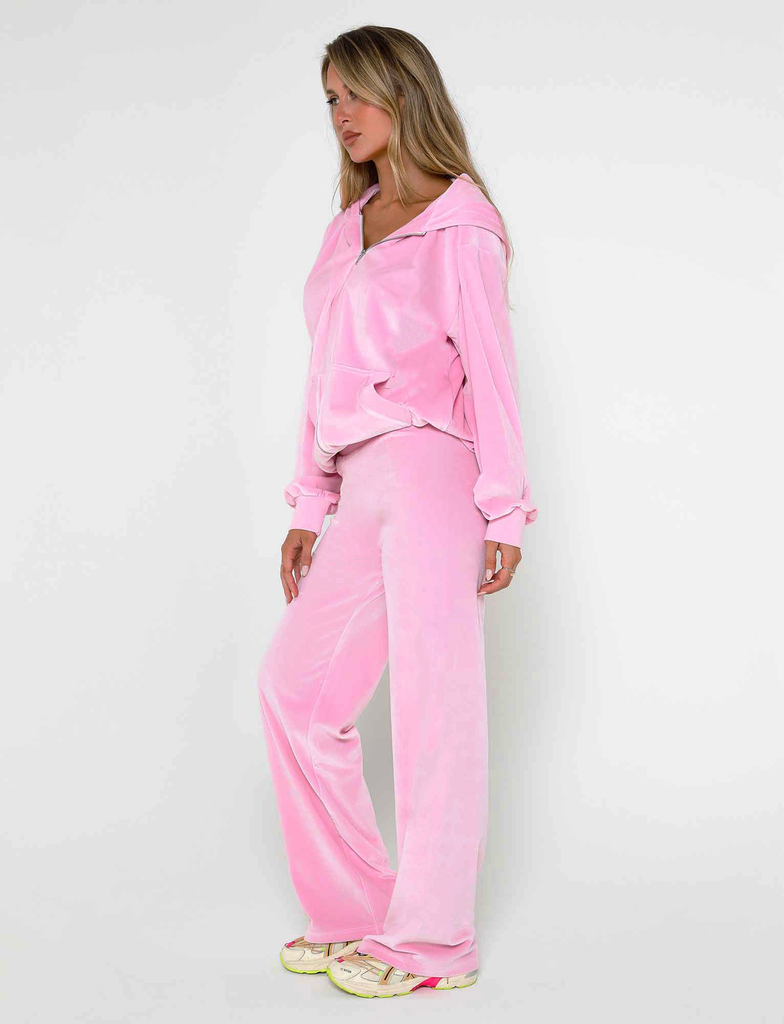 A woman stands against a plain white background, wearing the BABY PINK ERICKA PANT from TIGERMIST, part of a velour tracksuit that includes a loose-fitting hoodie and wide-leg pants. She has long blonde hair and stands in profile with her hands at her sides, accessorized with white and pink sneakers.