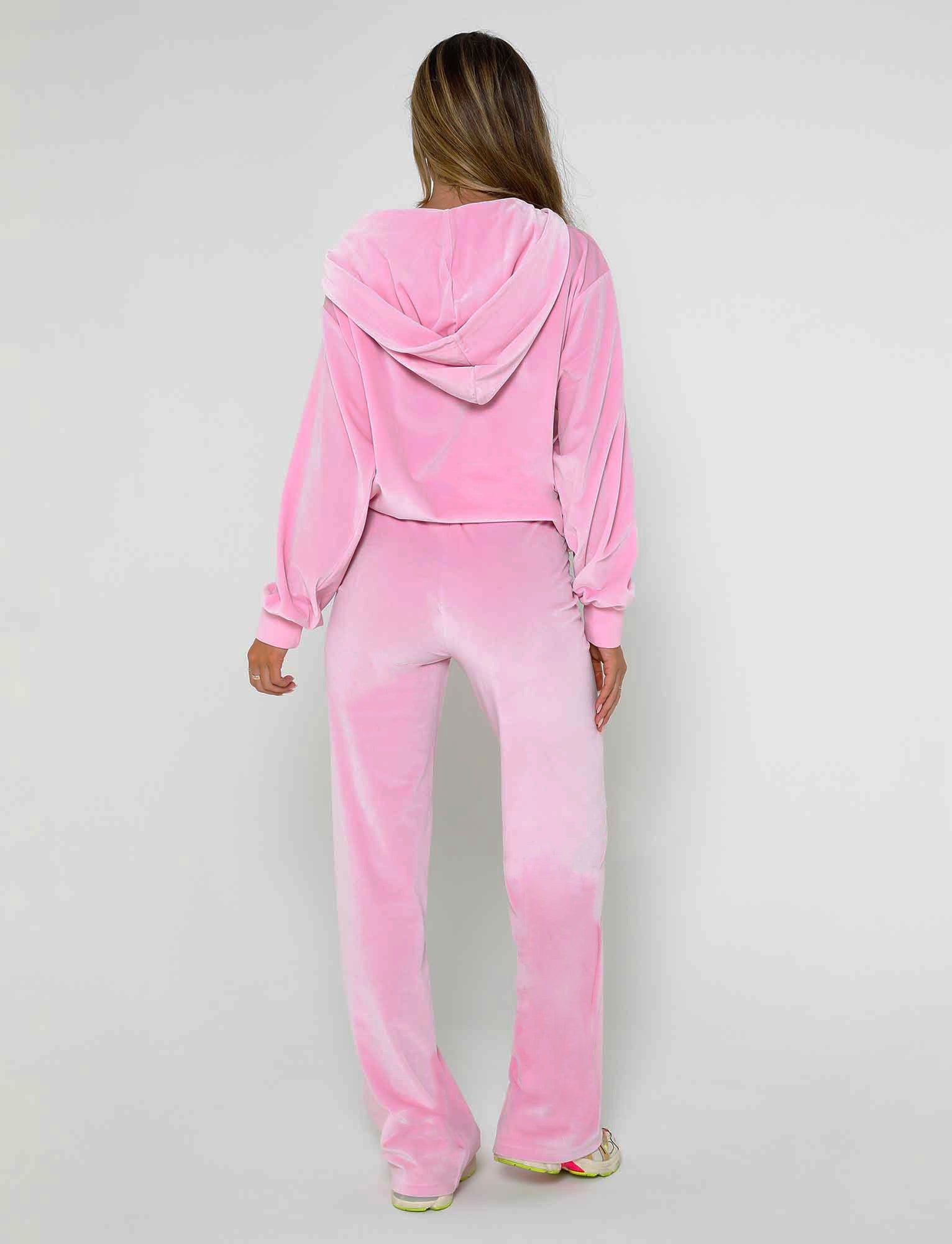A person with long hair is standing and facing away from the camera, wearing a TIGERMIST pink velour tracksuit. The outfit includes a hooded top and the ERICKA PANT in baby pink. They are also wearing light-colored sneakers. The background is plain and light-colored.