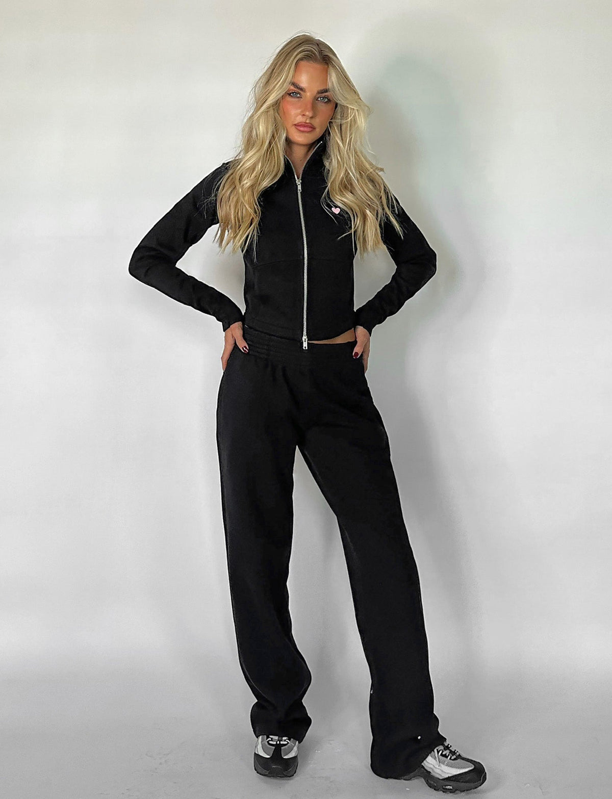 A person with long blonde hair stands against a white background, wearing a sleek Tiger Mist black zip-up jacket and matching VERDA PANT - BLACK from TIGERMIST. They are posing with their hands on their hips and sporting stylish black and white sneakers.