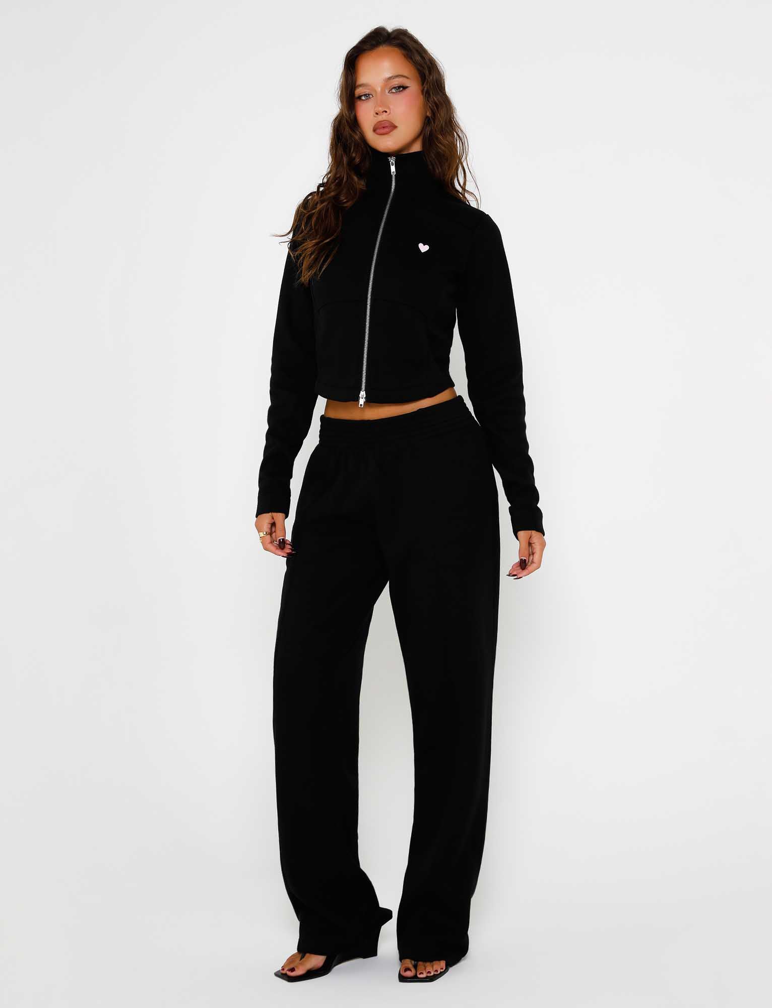 A woman stands against a plain white background, wearing the VERDA PANT - BLACK from TIGERMIST. She showcases a stylish black Tiger Mist outfit that includes a cropped zip-up jacket featuring a small heart detail paired with high-waisted wide-leg pants. With her long, wavy brown hair cascading down, she strikes a pose with one hand on her hip.