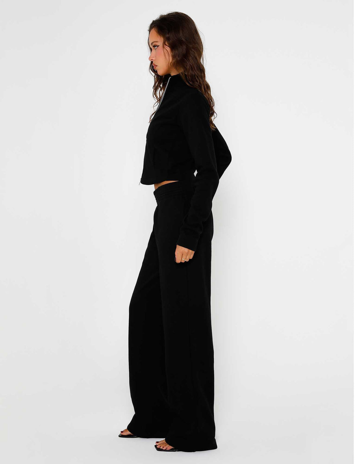 A woman with long, wavy hair stands sideways against a plain white background. She is wearing a black, long-sleeve crop top and the VERDA PANT - BLACK from TIGERMIST. She is looking forward, and her hands are relaxed by her sides.