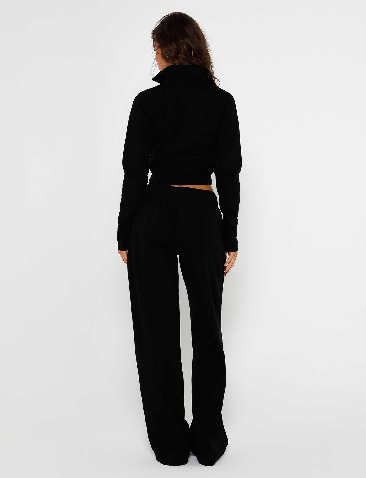 A person with long hair stands facing away from the camera, dressed in a sleek black long-sleeved top and the VERDA PANT - BLACK from TIGERMIST. The background is plain white.