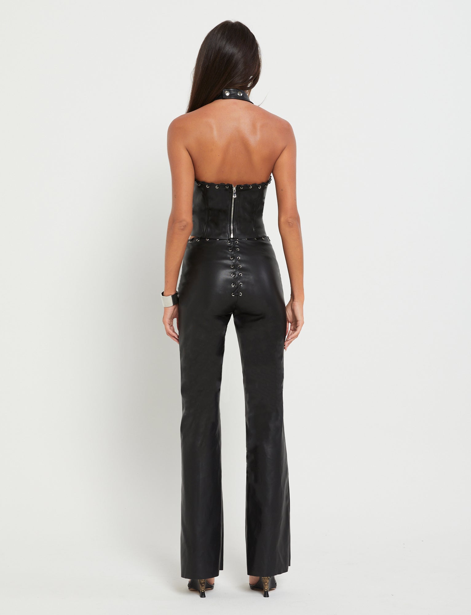 A person with long dark hair stands facing away from the camera, showcasing a stylish TIGERMIST BOSCO TOP in black PU and matching high-waisted pants with button details on the back. The background is plain white.