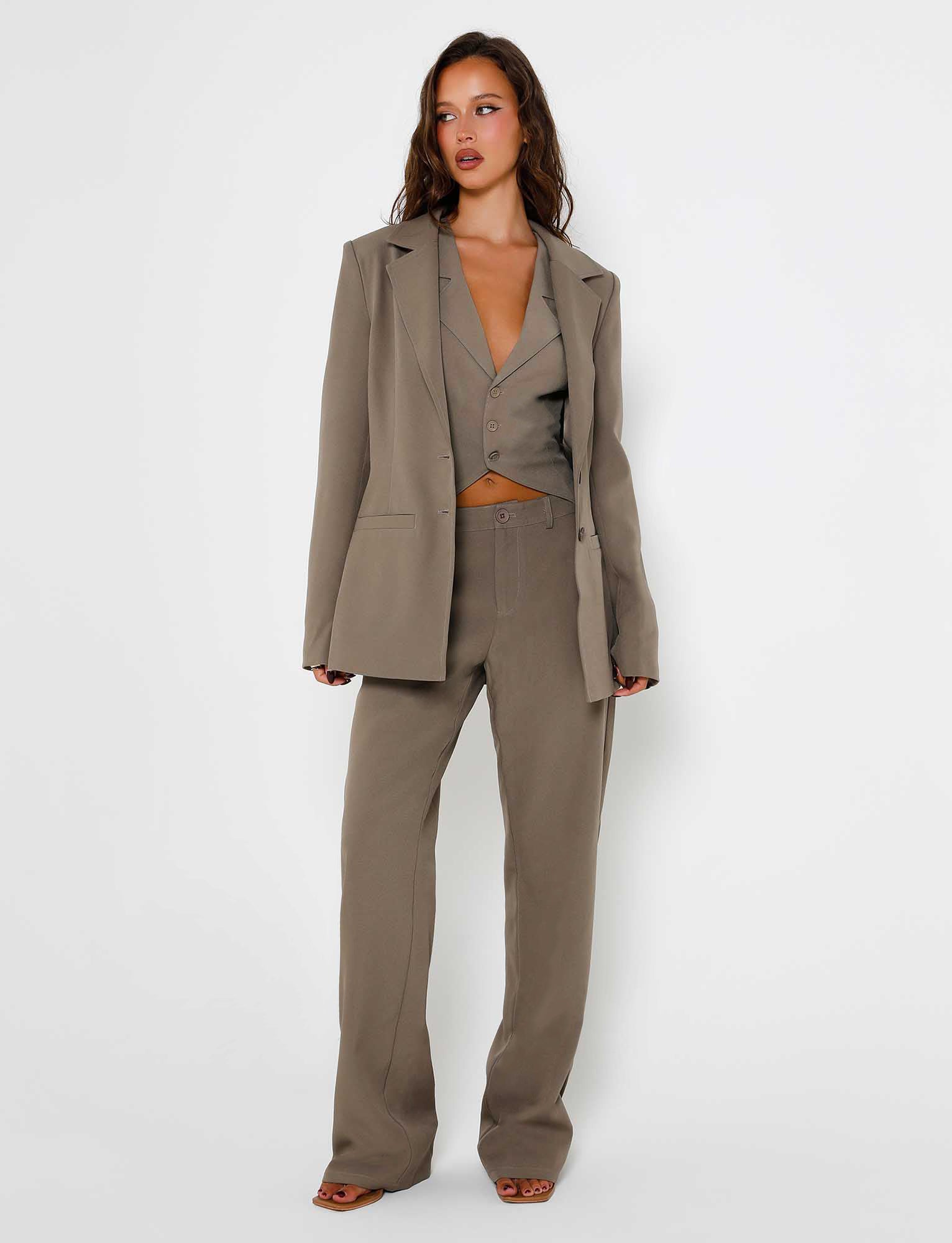 A woman stands against a plain white background, wearing a moss green pantsuit from TIGERMIST, featuring the Marjorie Jacket in Moss Green Khaki. She has long, wavy brown hair and is posing with her arms relaxed by her sides, looking slightly to the left.