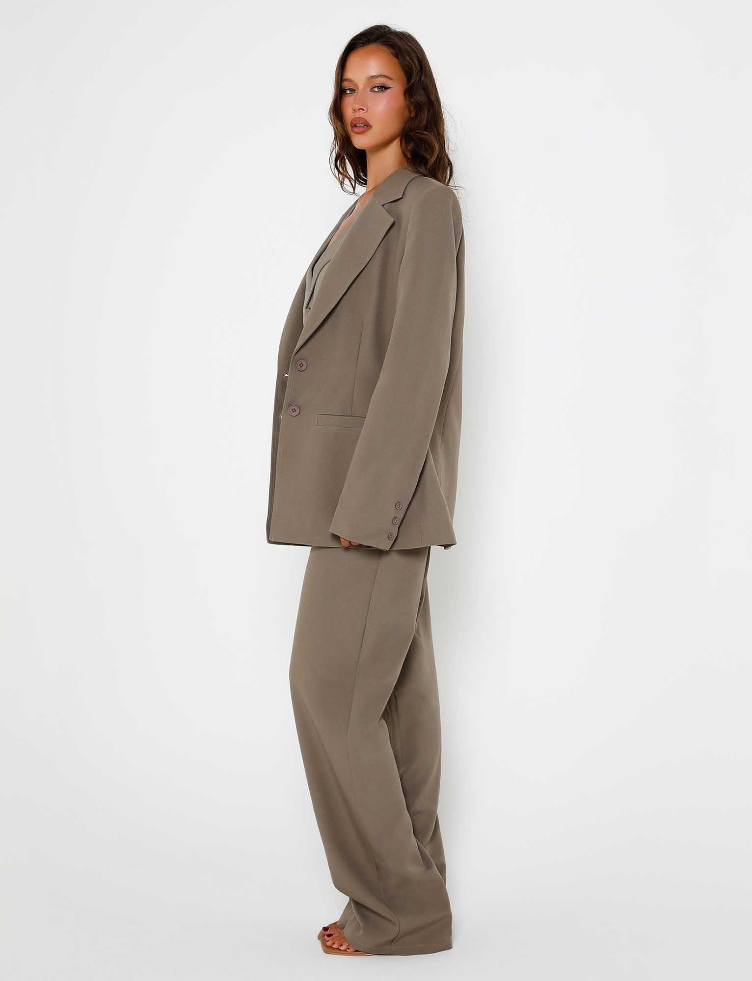 A woman with long, wavy hair stands against a plain white background, wearing an oversized khaki pantsuit featuring the MARJORIE JACKET in moss green from TIGERMIST. She is facing the camera at a slight angle, showing a side profile.
