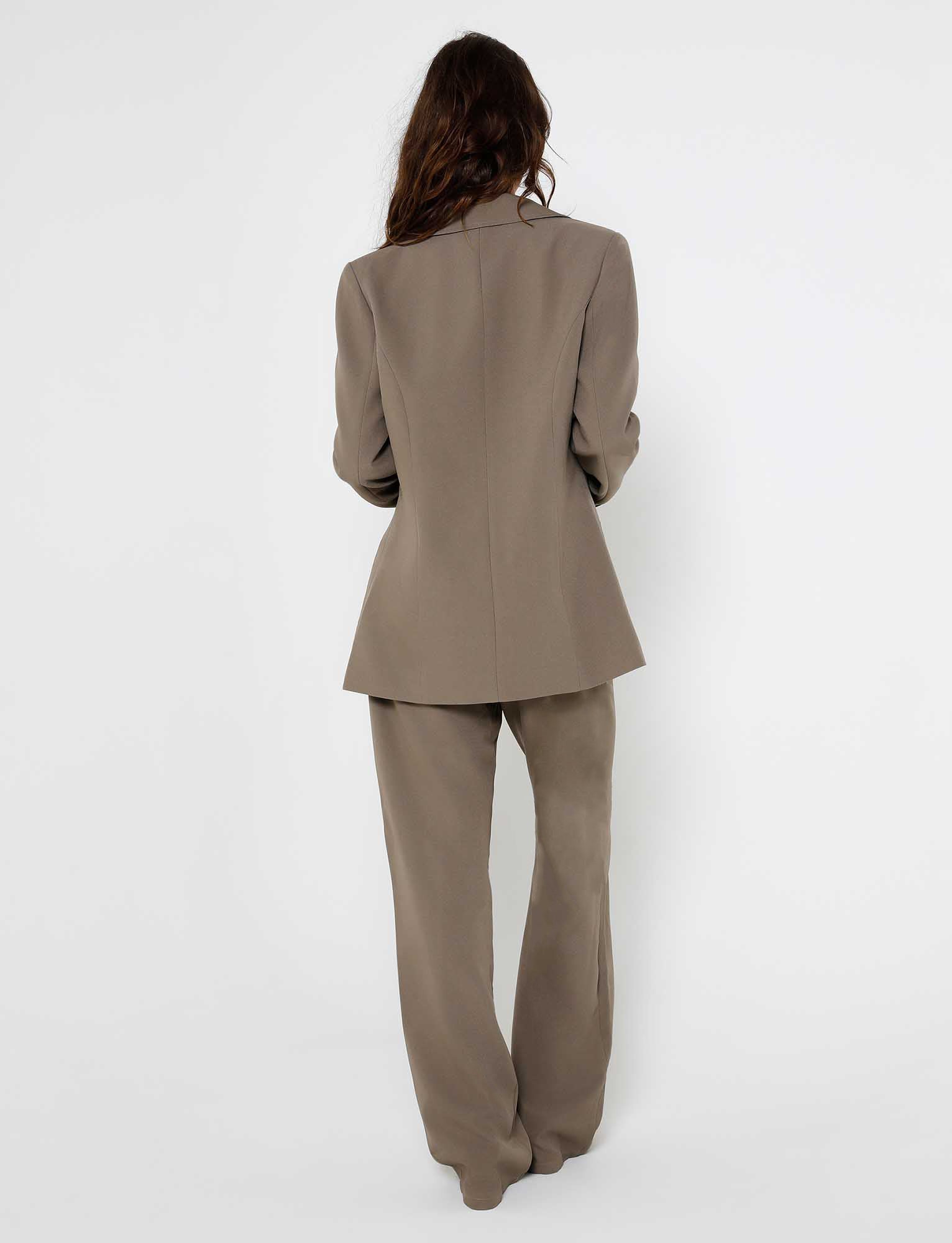 A person with long hair is standing with their back to the camera, wearing the MARJORIE JACKET from TIGERMIST in moss green and khaki. The jacket, paired with matching wide-leg pants, perfectly showcases the brand's flair for modern elegance against a plain white background.