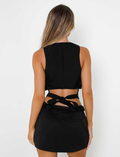 A woman with long hair is facing away from the camera, wearing the black MARJORIE VEST - BLACK and matching skirt from TIGERMIST. The back of her vest features criss-cross tie details at the waist. The background is white.