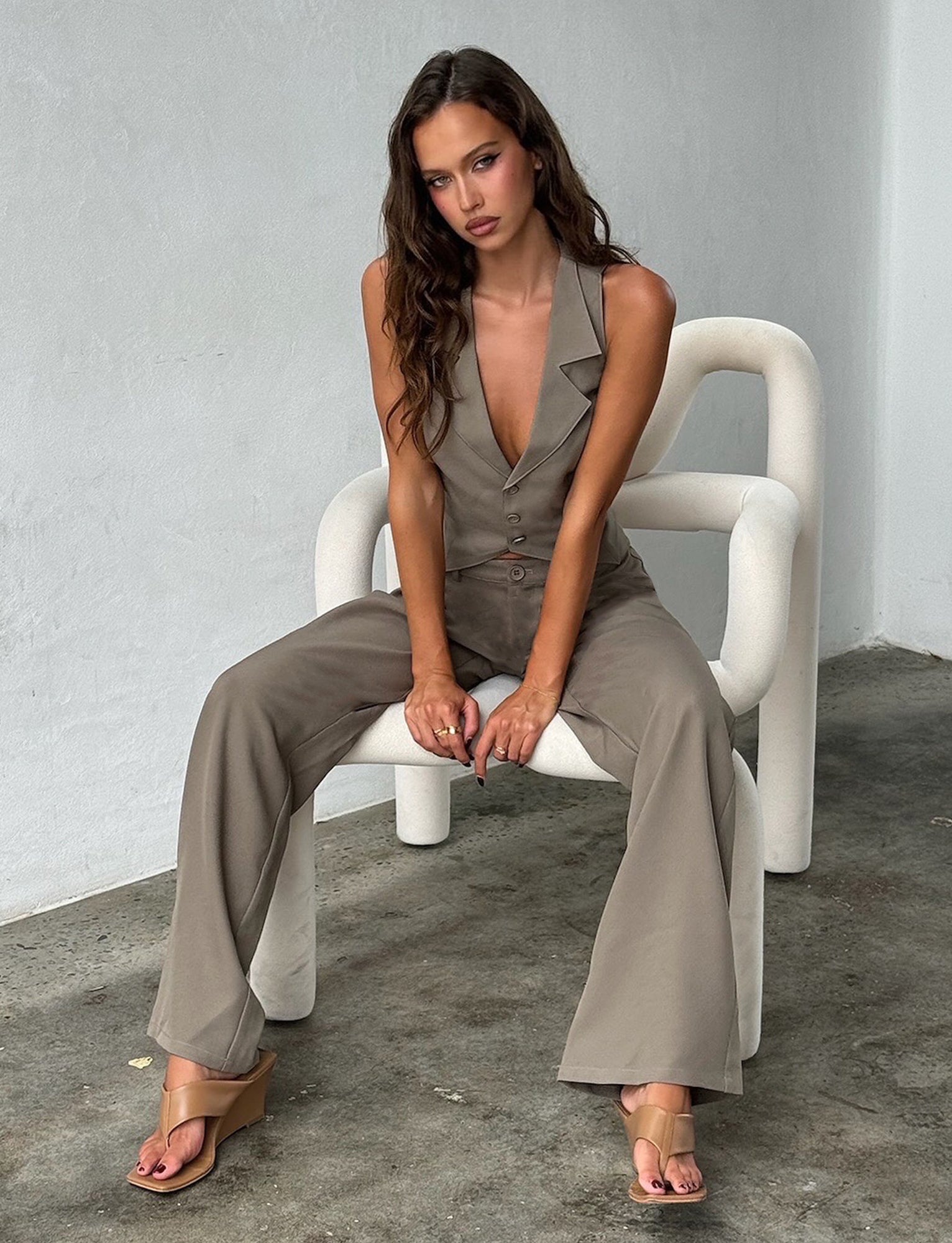 A woman with long, wavy dark hair is sitting on a modern, white chair. She is wearing the Marjorie Pant from Tiger Mist in moss green khaki, paired with beige sandals. The background is a minimalist indoor setting with white walls and a concrete floor.