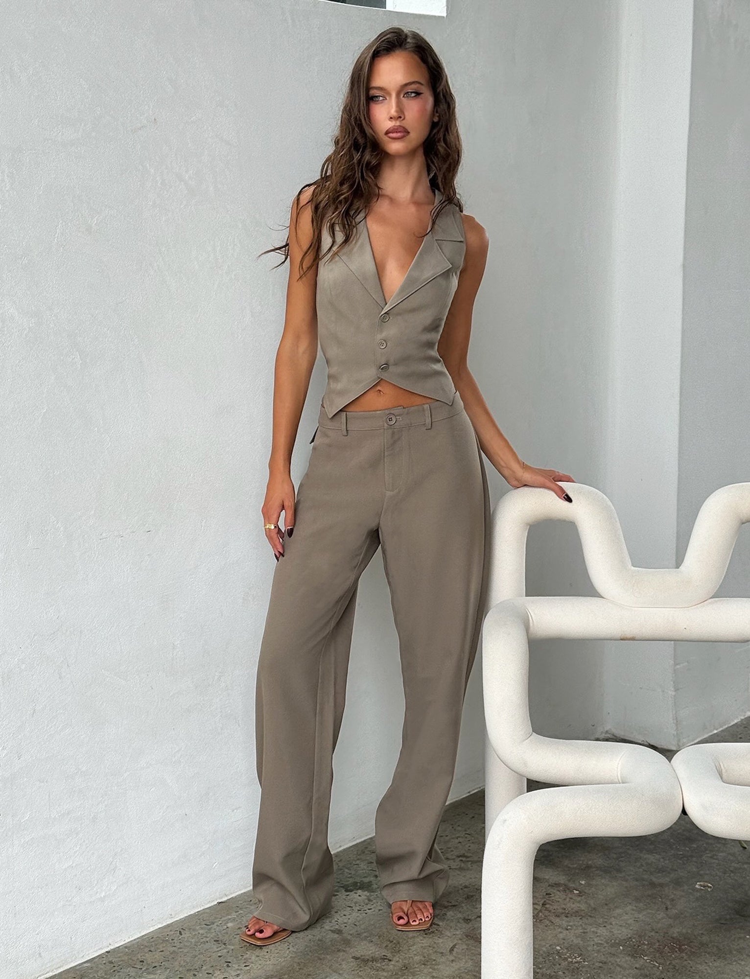 A woman with long, wavy hair wearing a tailored, sleeveless beige vest and the MARJORIE PANT in moss green Khaki from TIGERMIST stands beside a white, modernly designed chair in a minimalist room. She holds the chair with one hand, looking confidently at the camera.