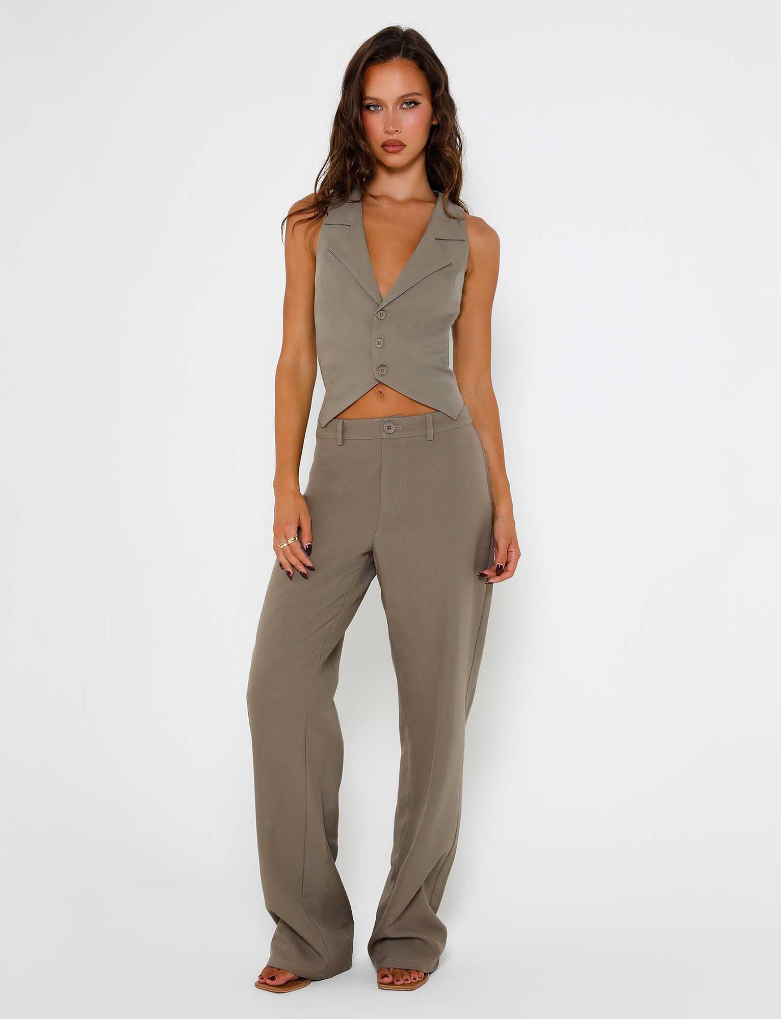 A woman with long brown hair stands against a plain white background, embodying the sleek elegance of TIGERMIST. She is wearing a sleeveless, button-up vest and the MARJORIE PANT in moss green khaki. Her left hand rests on her thigh while her right hand hangs by her side. She is expressionless, directly facing the camera.