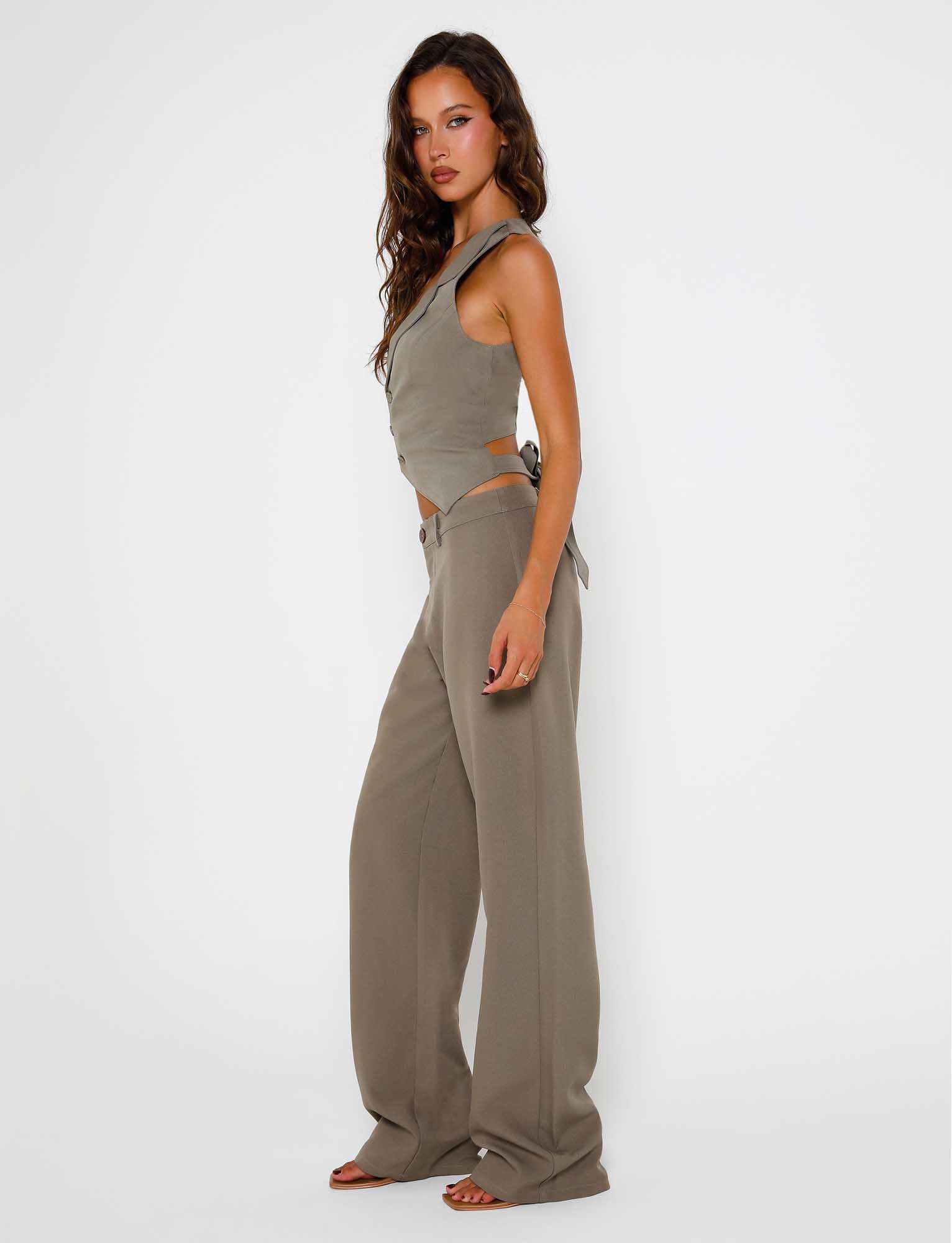 A woman stands against a plain white background wearing the MARJORIE PANT in moss green from the TIGERMIST collection. She has long, wavy brown hair and is looking directly at the camera with a neutral expression. She is barefoot and pairing her trousers with a gray sleeveless top featuring side cutouts.