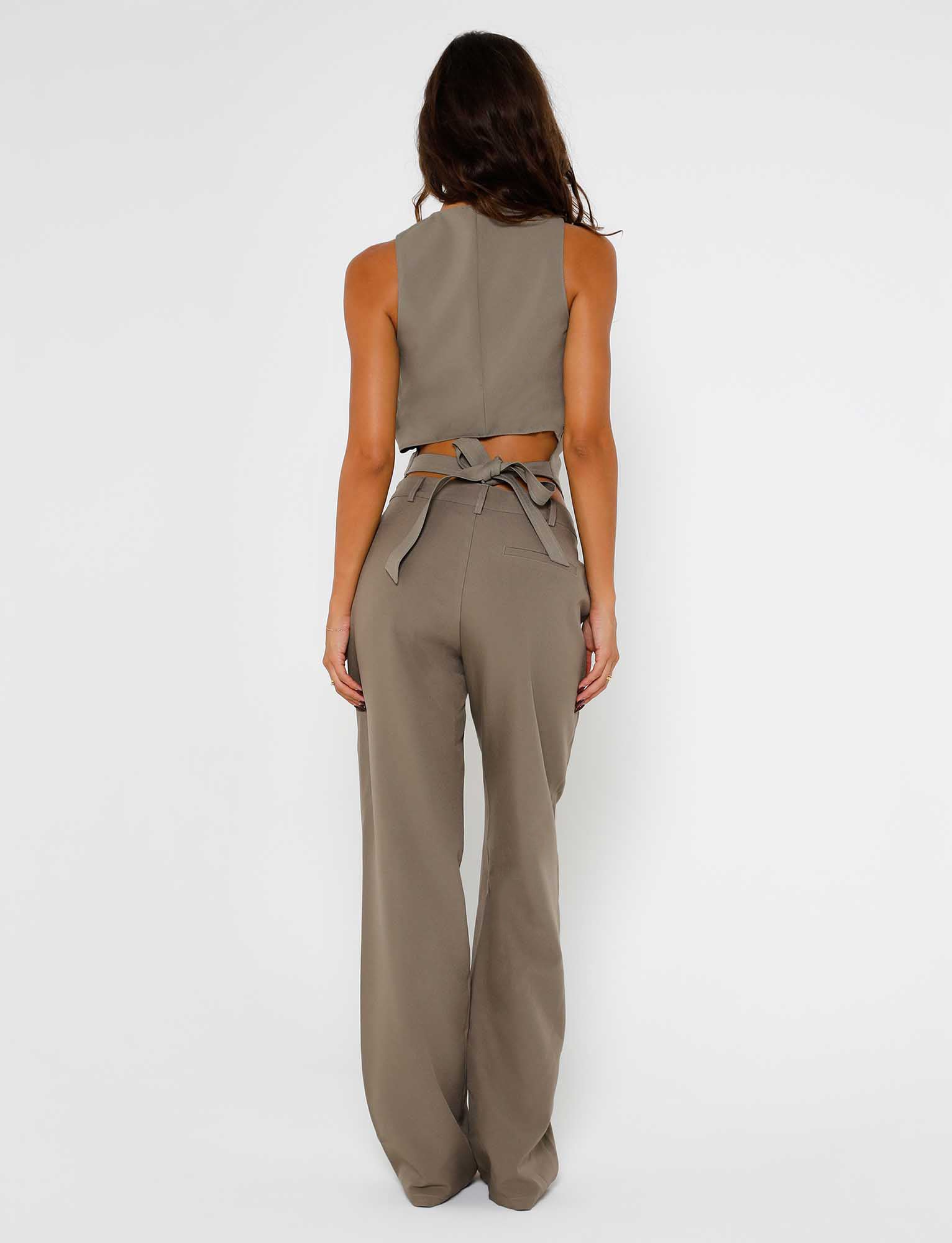 A person with long dark hair stands facing away, showcasing a coordinated set from Tiger Mist. They are wearing a grey sleeveless crop top featuring a back tie detail and the Marjorie Pant in moss green, which includes high-waisted wide-leg trousers accented with a belt tied in a bow. The background is plain white.