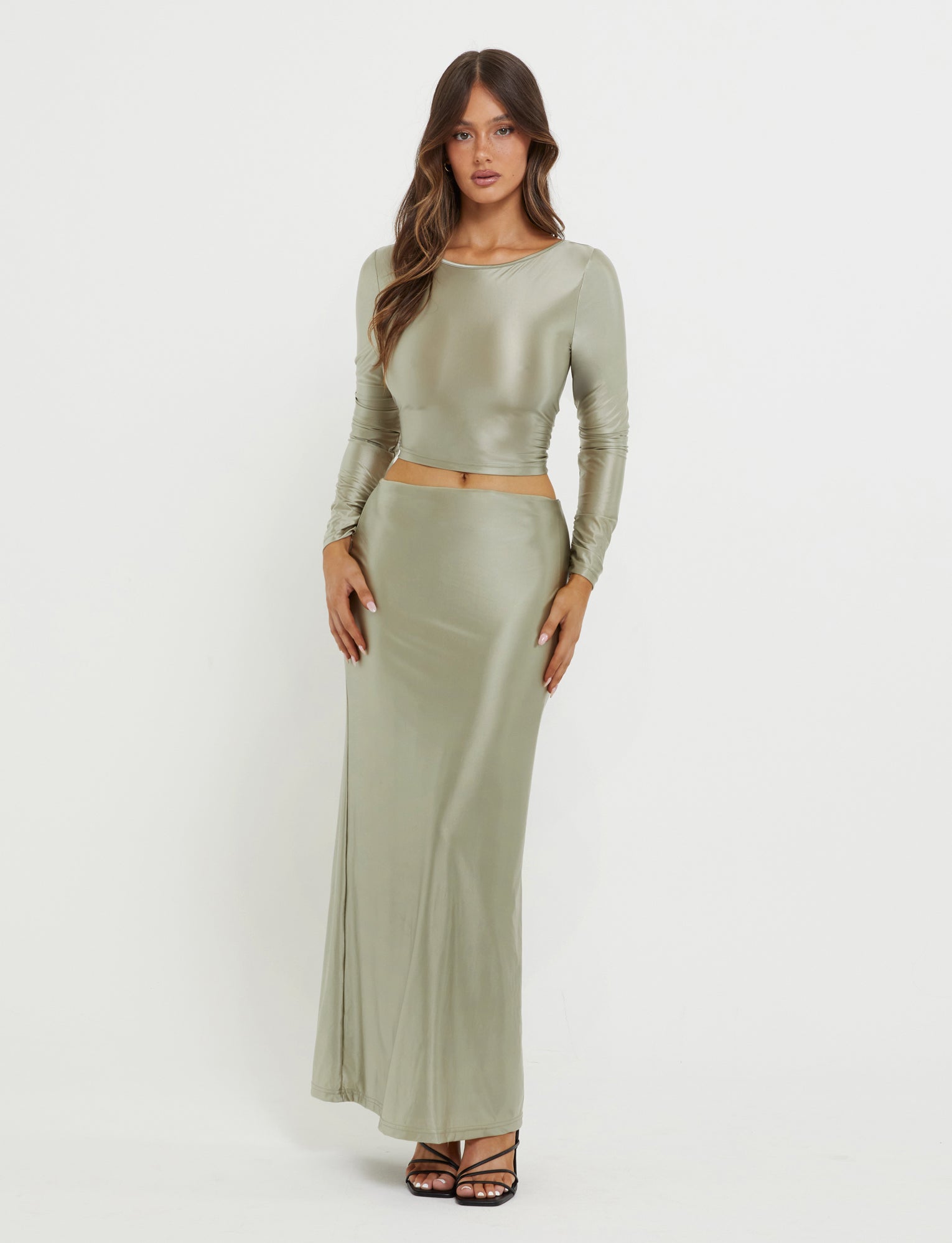 A woman stands against a plain white background wearing the form-fitting, long-sleeve THESSALY TOP in Matcha Green from TIGERMIST, paired with a matching high-waisted, ankle-length skirt. She is posing with both hands resting on her hips and is wearing strappy black high heels.