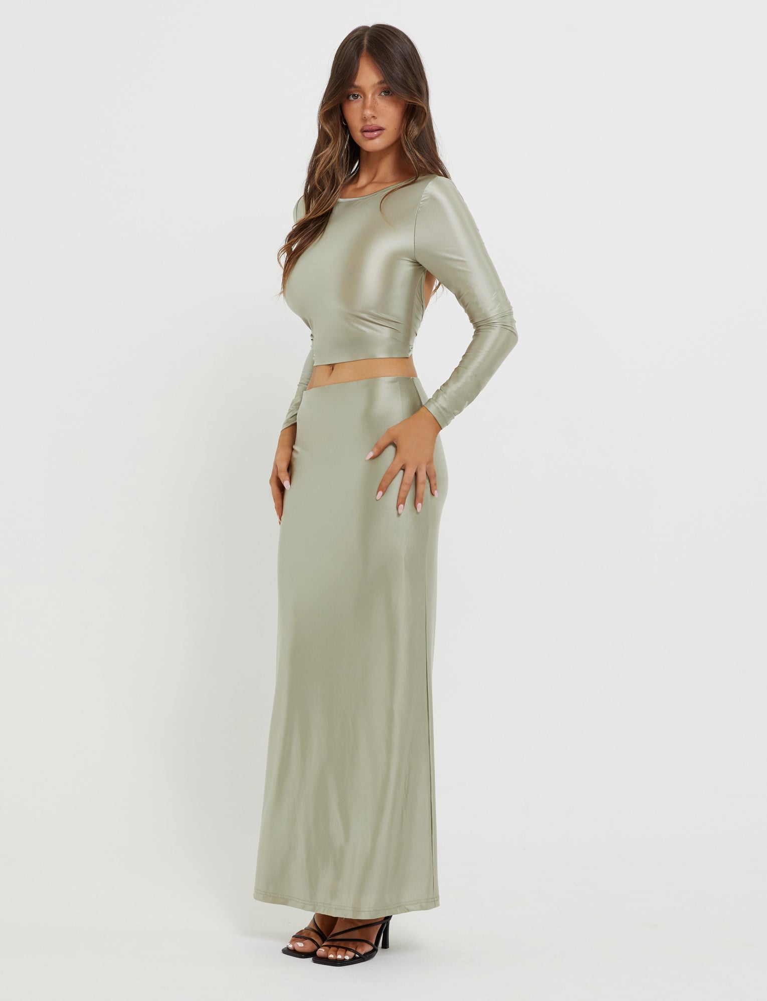 A woman with long, wavy hair is wearing the form-fitting Thessaly Top in Matcha Green by TIGERMIST, which features long sleeves and a cutout at the waist. She pairs it with a metallic olive-green maxi skirt and black high-heeled sandals. She stands sideways with one hand on her hip against a white background.