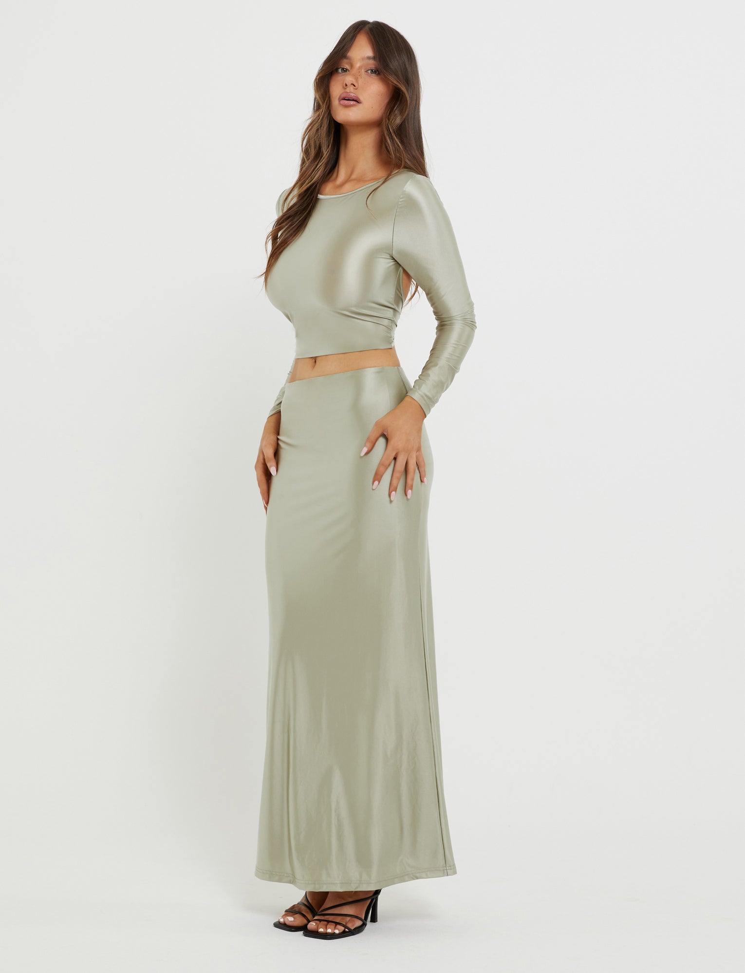 A woman with long brown hair is wearing a shiny, form-fitting, Matcha Green Thessaly Top from Tiger Mist. She pairs it with a coordinating high-waisted, floor-length skirt and black high-heeled sandals. She stands against a plain white background with one hand on her hip.