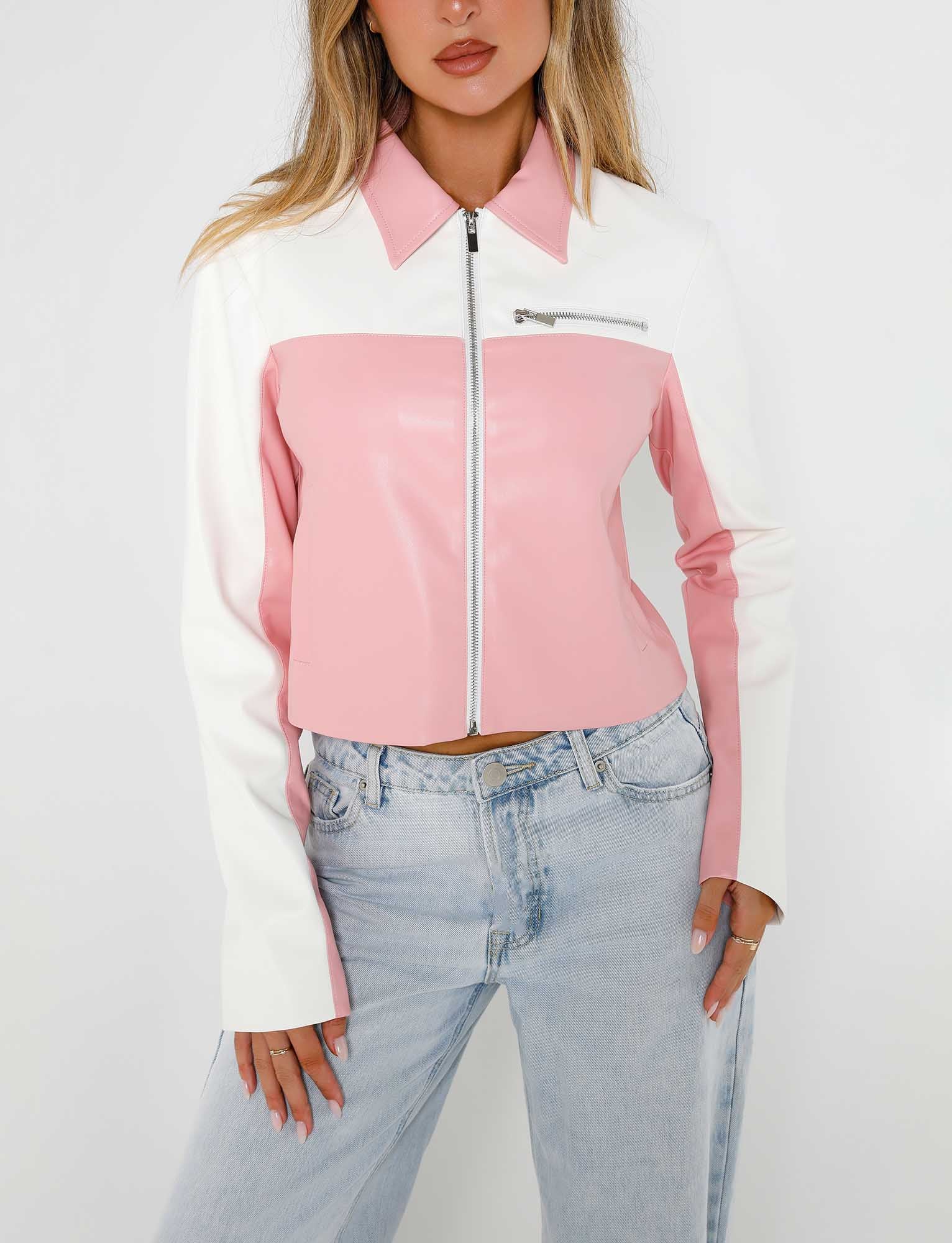 A person stands against a plain background wearing the TIGERMIST DELIA JACKET - PINK, which features a two-toned design with a white top half and pink bottom half. The jacket includes a front zipper and a zippered chest pocket. They are also wearing light blue jeans, with their hands resting in their pockets.