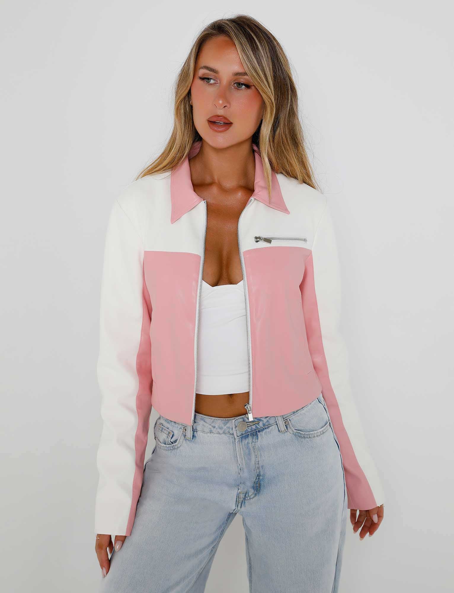 A woman with long blonde hair stands against a plain white background. Clad in a stylish TIGERMIST DELIA JACKET - PINK : PINK/WHITE with a front zipper over a white top, she pairs it with light blue high-waisted jeans. Her expression is neutral as she gazes slightly to the side.