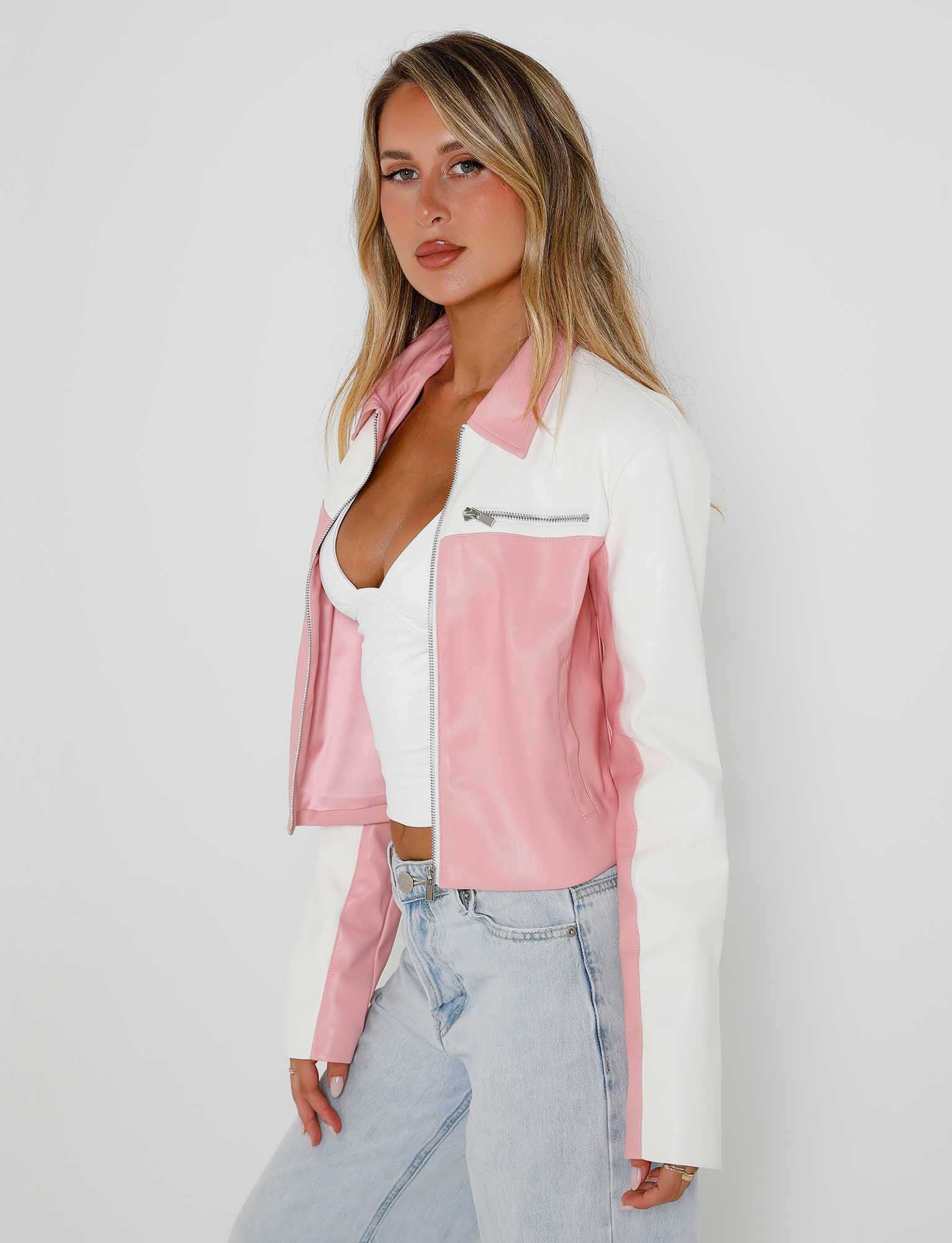 A woman with long blonde hair is wearing the TIGERMIST DELIA JACKET - PINK in a pink and white color scheme over a white top and light blue jeans. She is posing against a plain light background, with her right hand on her hip, exuding confidence as she looks at the camera.